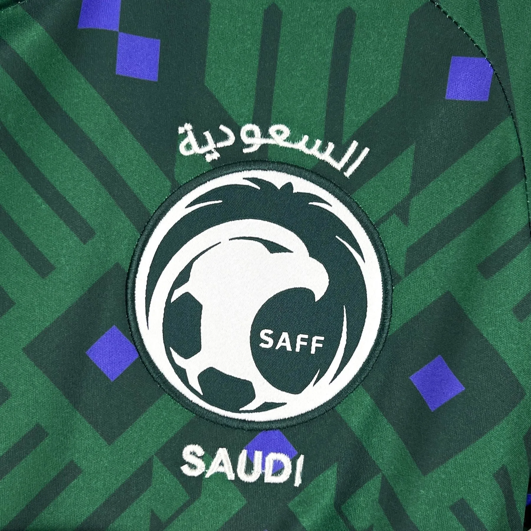 Escudo oficial de la SAFF (Saudi Arabian Football Federation) bordado en blanco y negro sobre el pecho de la camiseta verde del Mundial 2026.