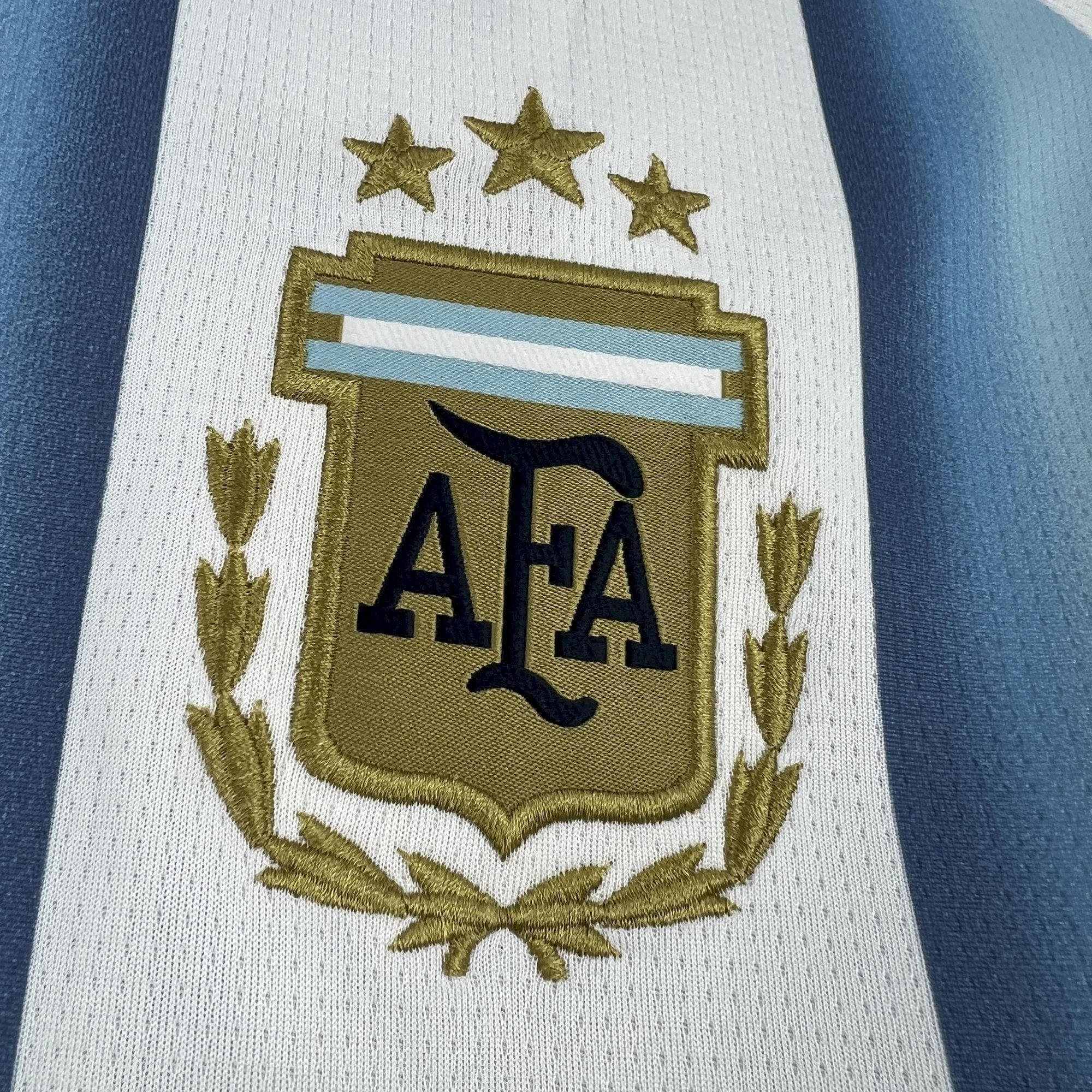 Detalle en primer plano del escudo bordado de la Asociación del Fútbol Argentino (AFA) con las tres estrellas doradas y laureles sobre las franjas celestes y blancas.