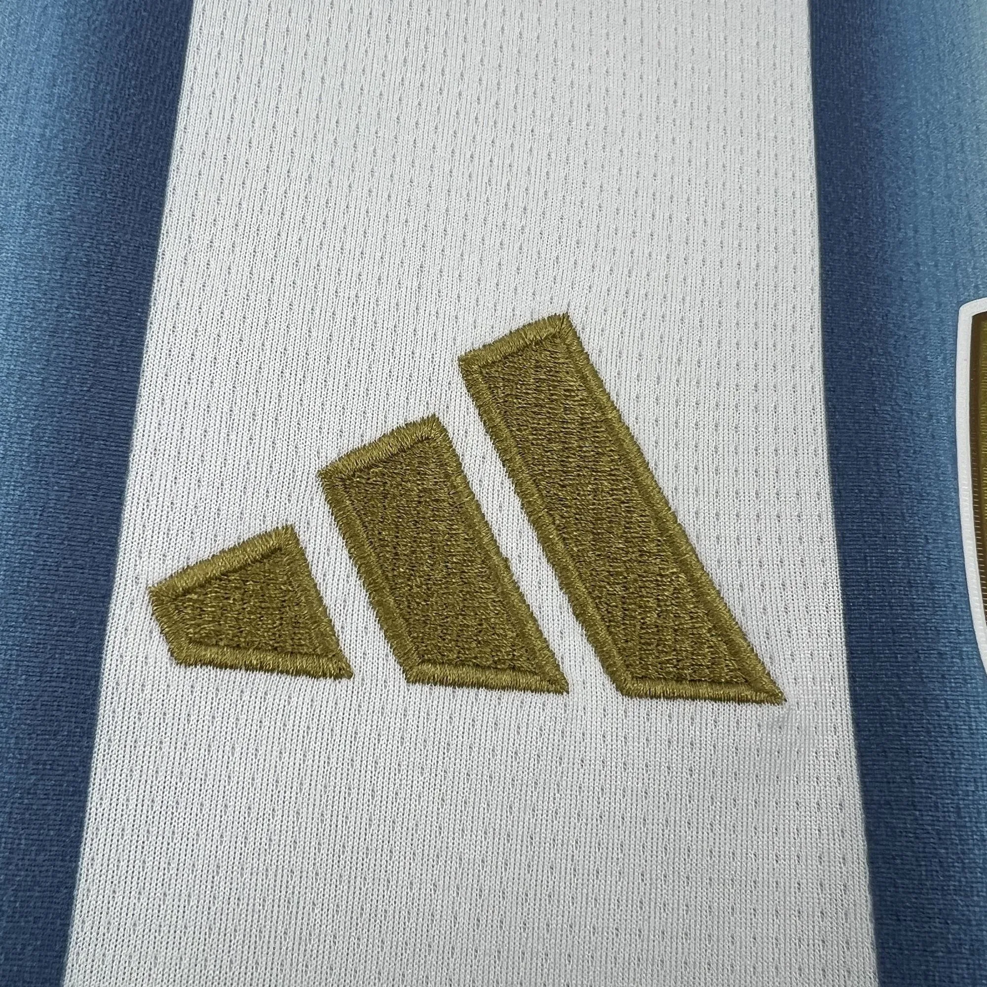 Primer plano del logotipo de la marca bordado con hilo dorado de alta densidad sobre una de las franjas blancas del tejido técnico de la camiseta de Argentina.