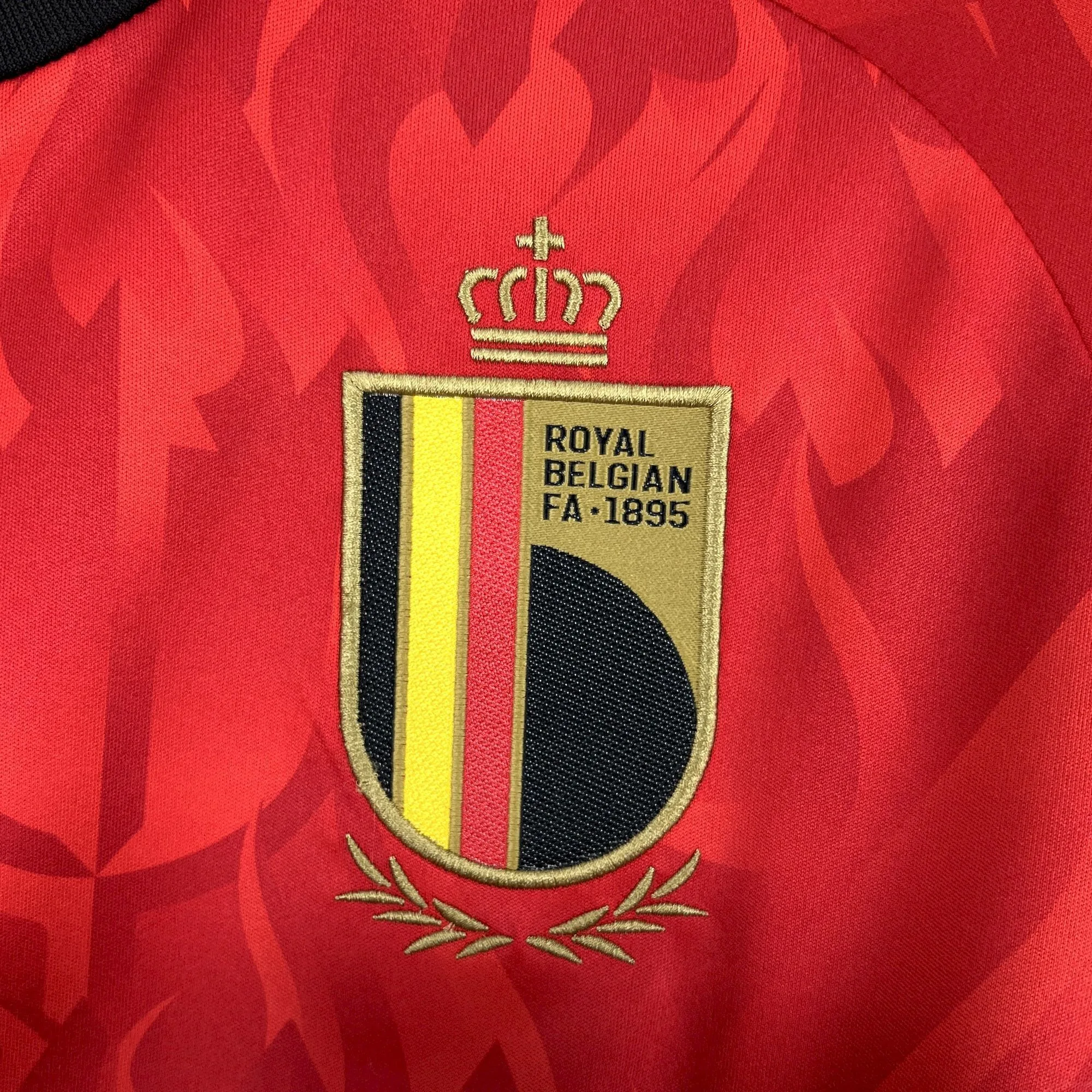 Primer plano del escudo de la Real Federación Belga de Fútbol bordado sobre fondo rojo con motivos geométricos.