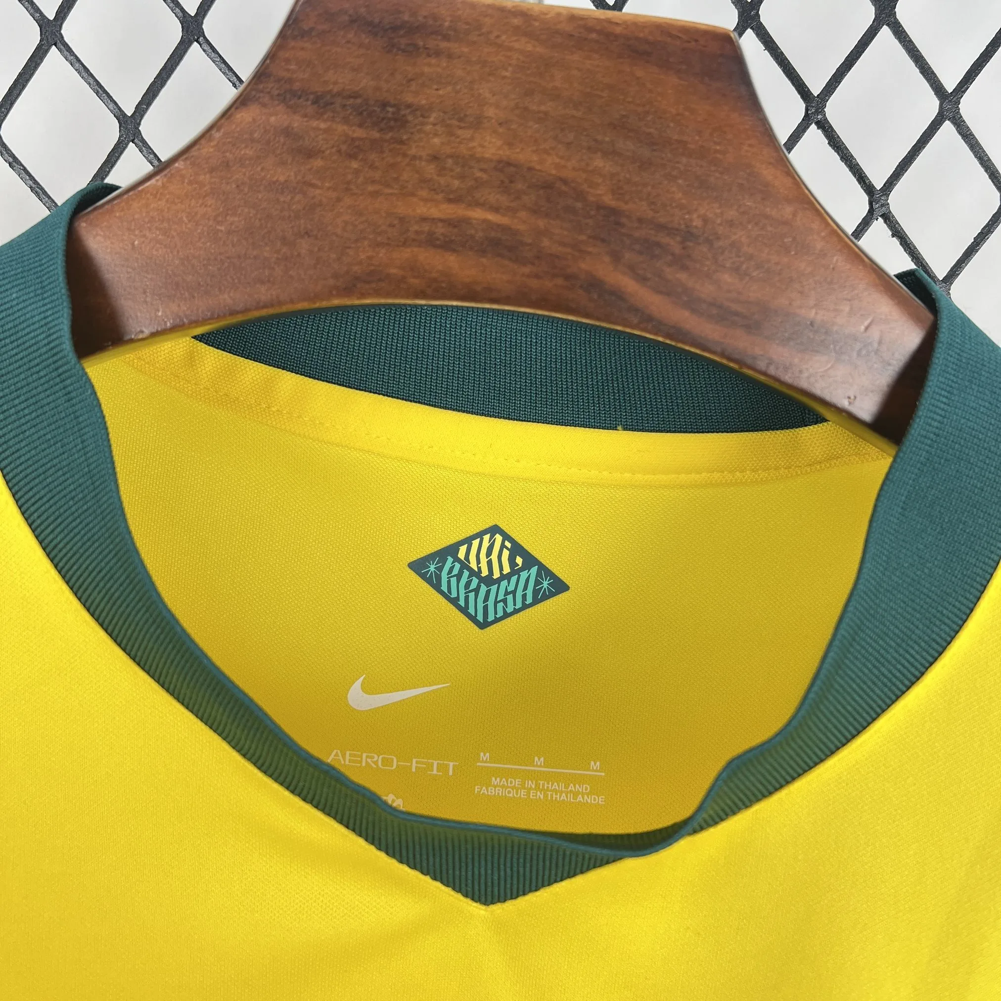 Vista de cerca del cuello verde de la camiseta de Brasil. En el interior amarillo se aprecia un sello en forma de diamante con la palabra "BRASIL" en estilo gráfico, el logotipo de la marca, la tecnología "AERO-FIT", la talla "M" y el sello de fabricación "MADE IN THAILAND".
