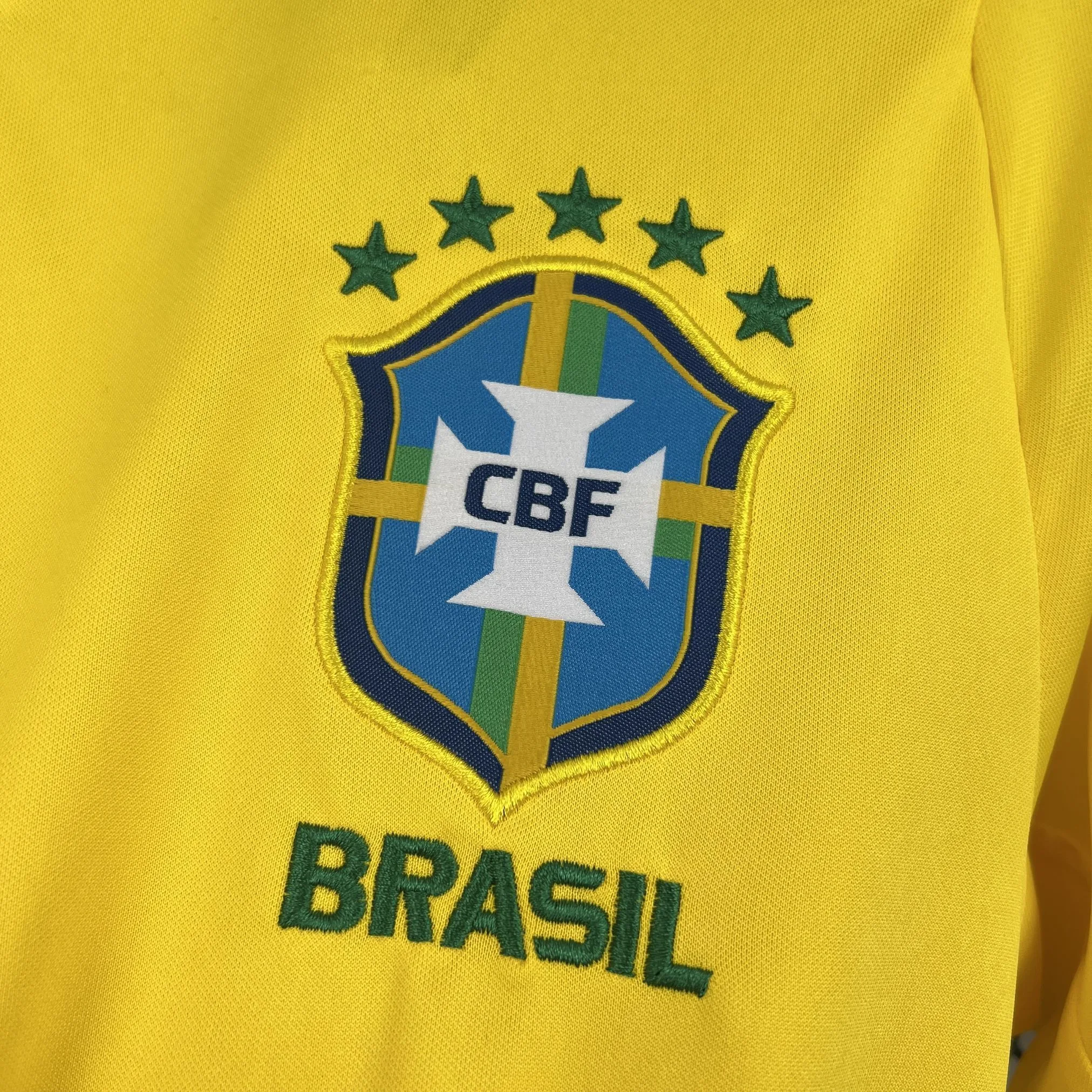 Primer plano del escudo de la Confederación Brasileña de Fútbol (CBF) bordado en el pecho. Presenta una cruz blanca central con las siglas "CBF" sobre un fondo azul y amarillo, coronado por cinco estrellas verdes que representan los títulos mundiales y con la palabra "BRASIL" bordada en verde en la parte inferior.