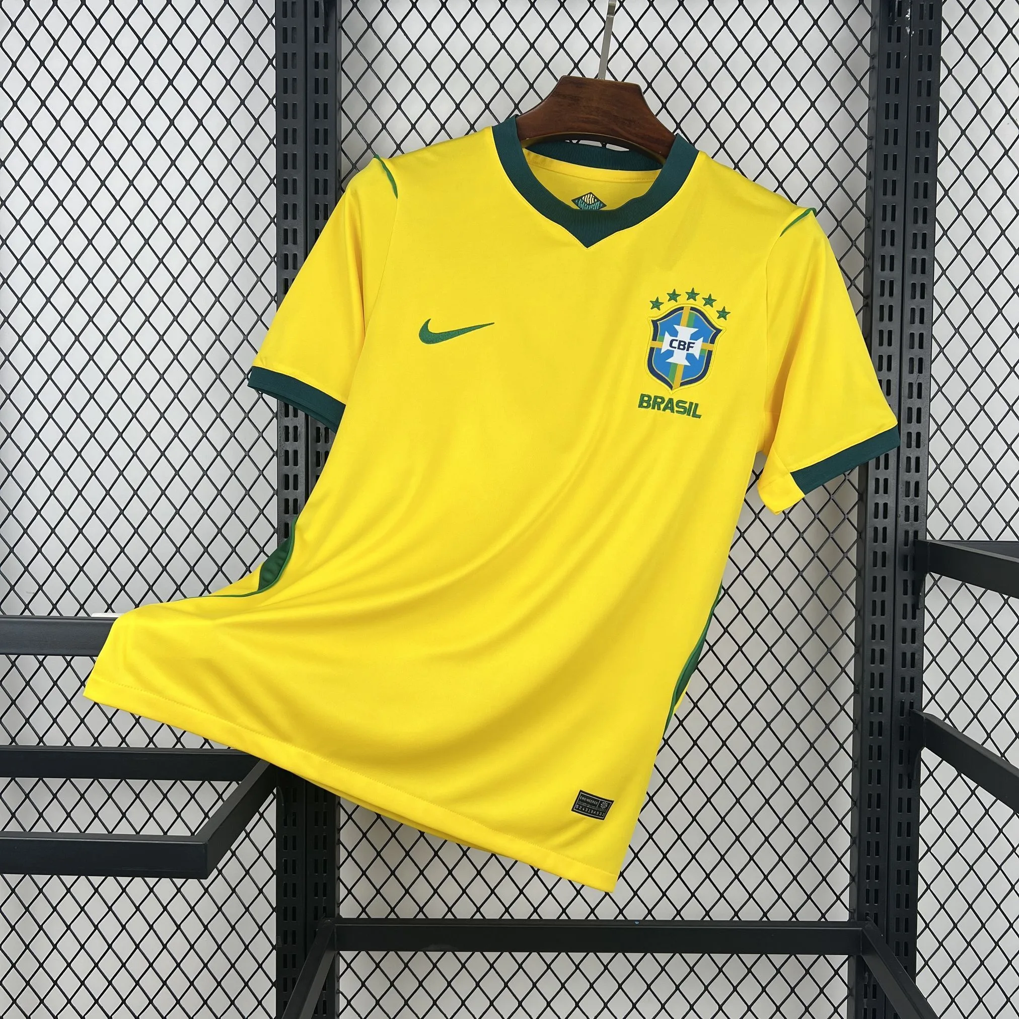 Camiseta de la selección de Brasil en su clásico color amarillo vibrante. Presenta un cuello redondo en color verde oscuro con un detalle de inserción triangular en la base, puños de las mangas también en verde y paneles laterales del mismo tono. En el pecho destaca el escudo de la Confederación Brasileña de Fútbol (CBF) con sus cinco estrellas verdes y el logotipo de la marca en color verde.