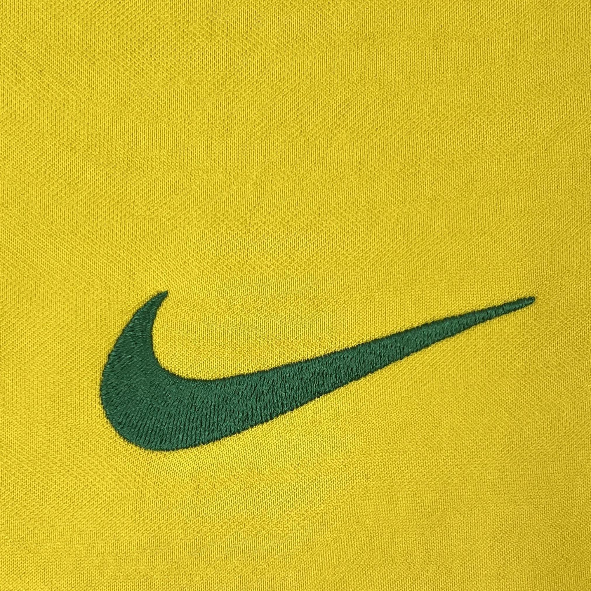 Primer plano del icónico logotipo de la marca deportiva bordado con hilo de color verde oscuro sobre el tejido amarillo de la camiseta de Brasil.