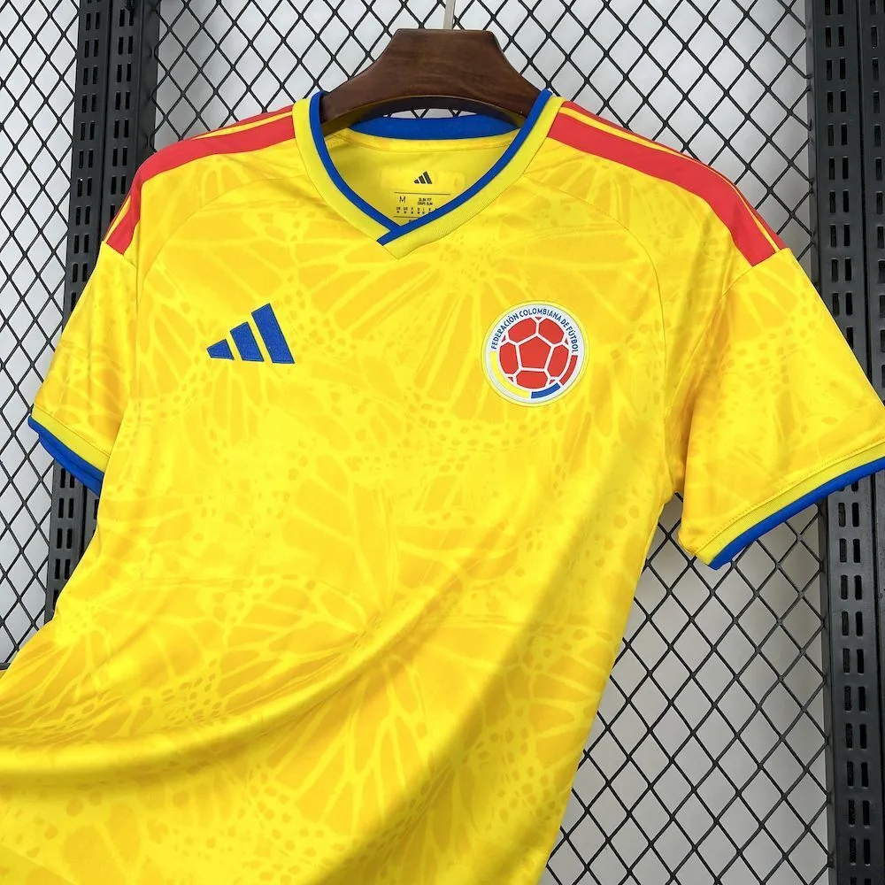 Vista frontal ladeado de la camiseta de Colombia para el Mundial 2026, en color amarillo tradicional con un sutil patrón de mariposas tonales y detalles en azul y rojo.