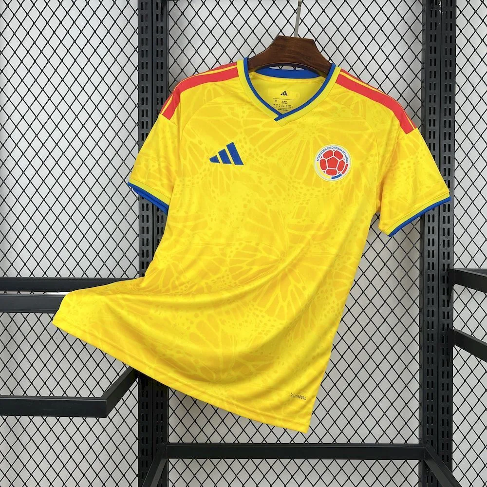 Vista frontal de la camiseta de Colombia en amarillo tradicional con un patrón de mariposas tonales y detalles en azul y rojo.