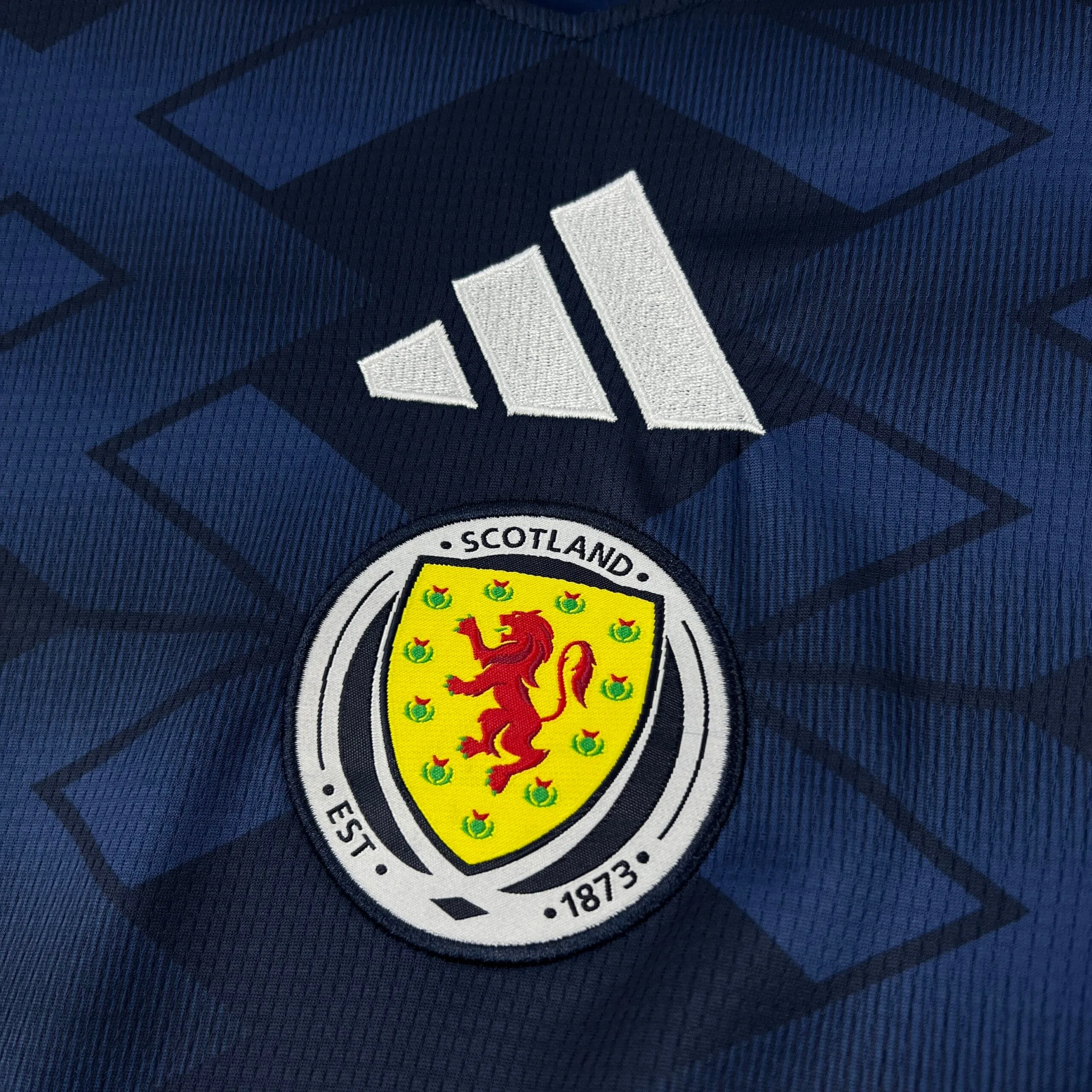 Primer plano del escudo circular de la selección de Escocia con el león rampante sobre fondo amarillo y el logotipo de la marca bordado en blanco sobre el tejido azul con patrón geométrico.