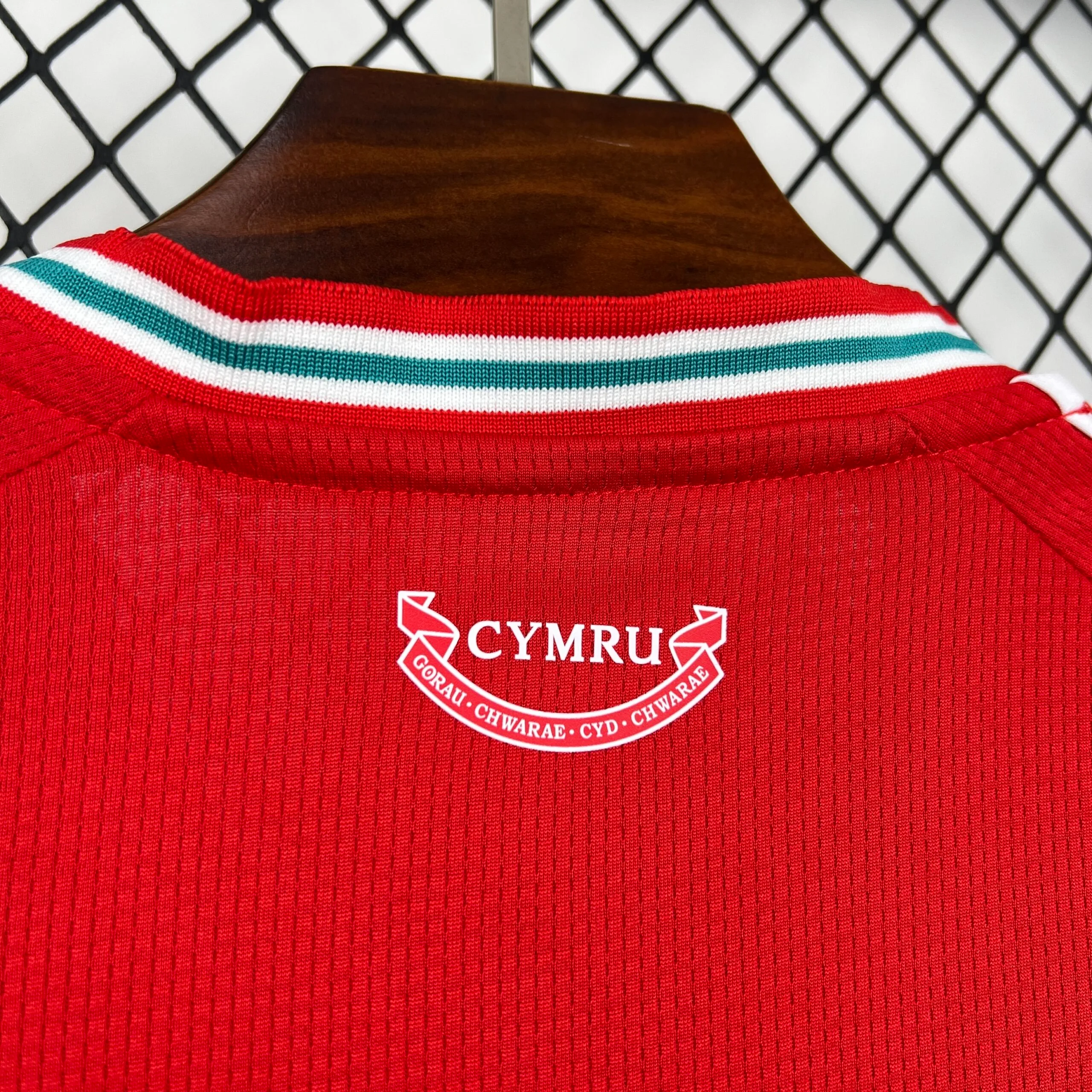 Primer plano de la parte posterior del cuello de la camiseta roja, que muestra un sello gráfico blanco con la palabra "CYMRU" y el lema en galés "GORAU CHWARAE CYD CHWARAE" dentro de una cinta, justo debajo del ribete tricolor.