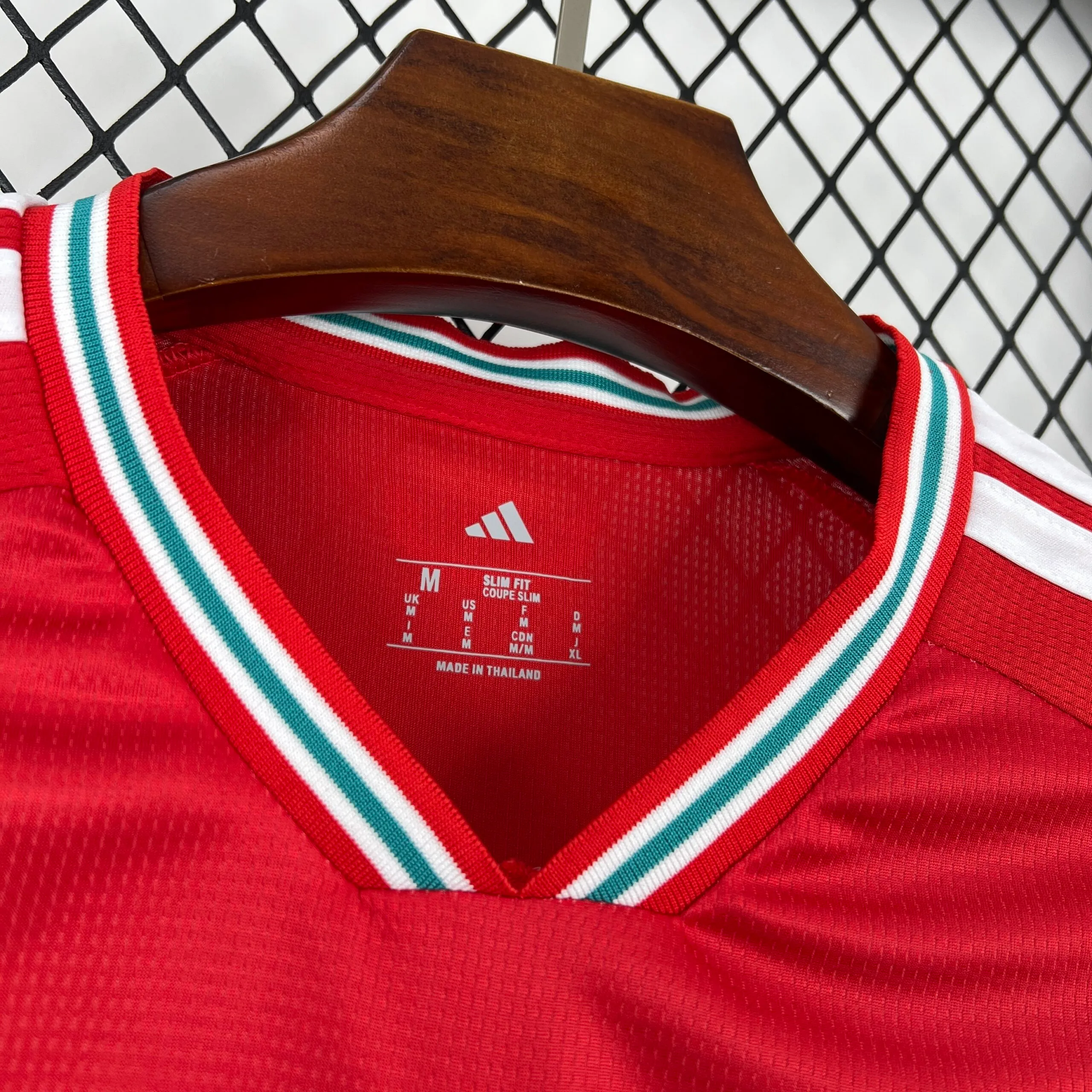 Primer plano del interior del cuello en V de la camiseta de la selección de Gales, que muestra un ribete elástico con rayas blancas, verdes y rojas. En el tejido rojo microperforado se aprecia el logotipo de la marca, la talla "M", el corte "SLIM FIT" y el sello de fabricación "MADE IN THAILAND" impresos en color blanco.