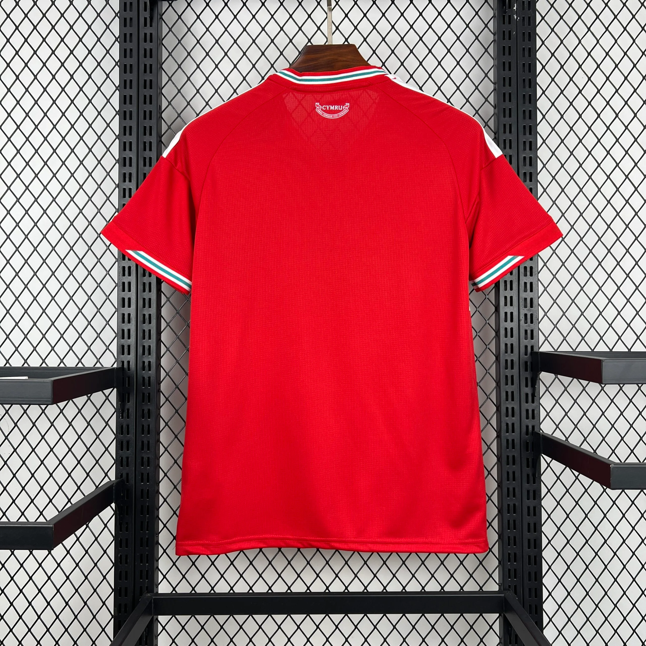 Parte posterior de la camiseta en color rojo sólido, que presenta un diseño limpio con las tres franjas blancas en los hombros. Destaca el ribete tricolor (blanco, verde y rojo) en el cuello y en el remate de las mangas, además de un pequeño sello con la palabra "CYMRU" y el diseño de un narciso en la zona de la nuca.
