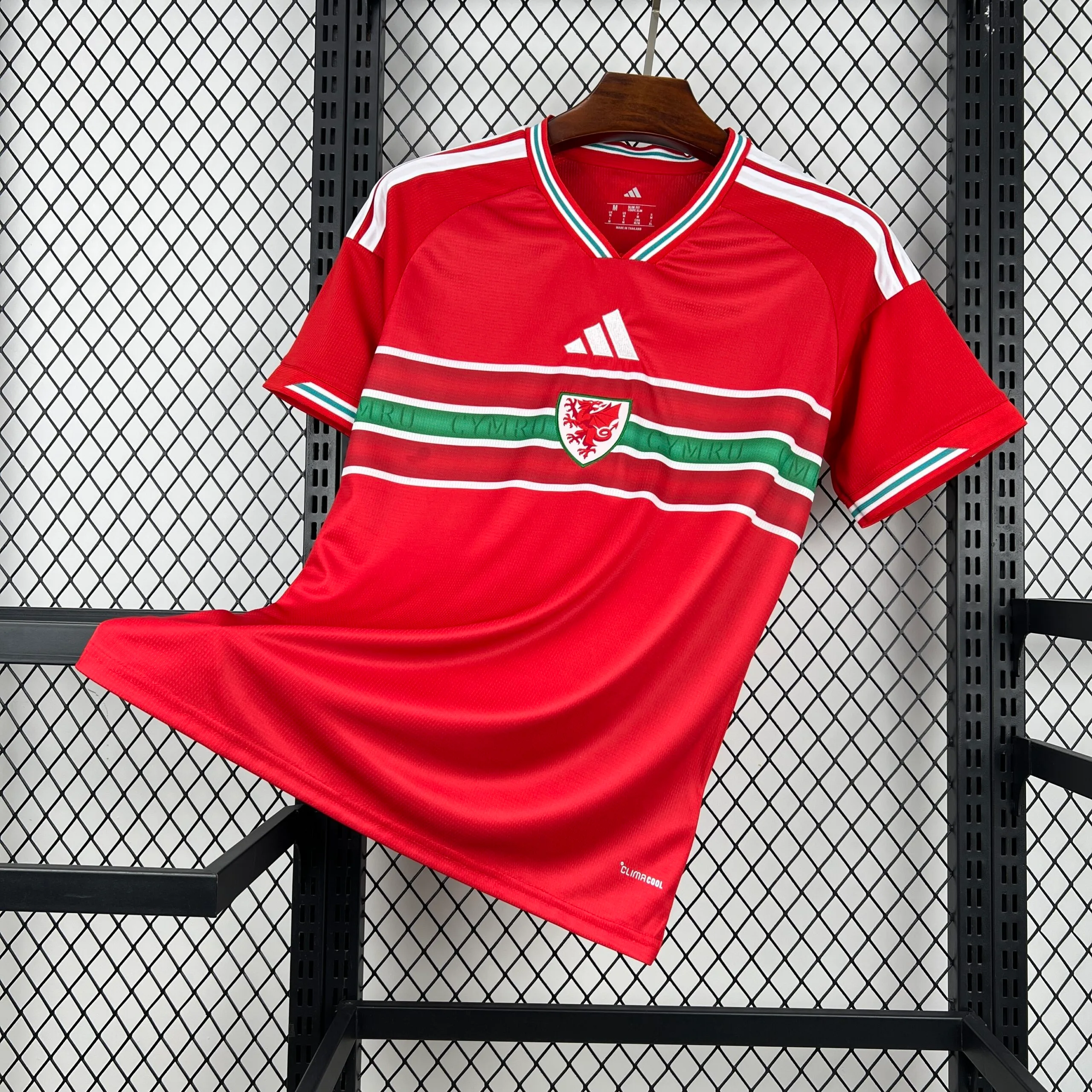 Vista frontal de la camiseta de la selección de Gales en color rojo, que presenta una franja horizontal tricolor (blanca, verde con la palabra "CYMRU" y blanca) en el pecho, donde se ubica el escudo del dragón. El diseño incluye cuello en V con ribetes multicolores, las tres franjas blancas en los hombros y el logotipo de la marca centrado sobre la franja.