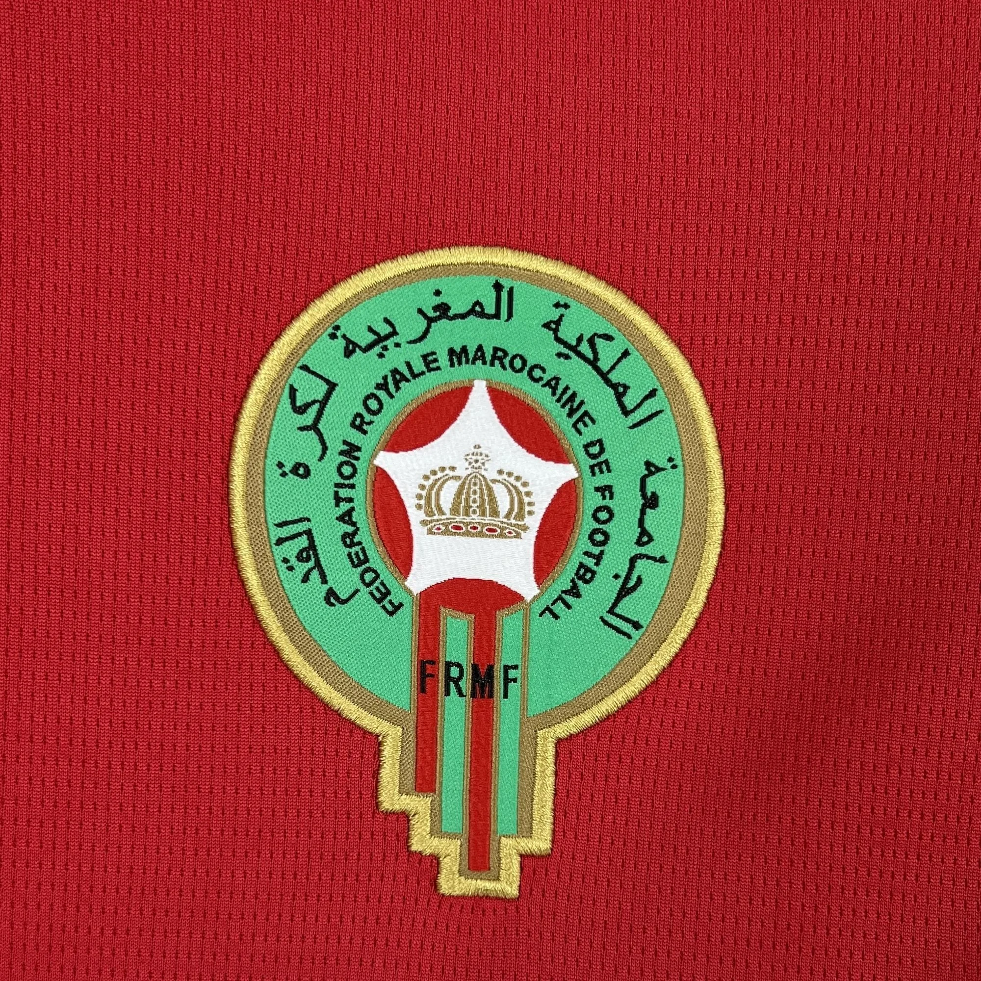 Primer plano del escudo bordado de la Federación Real Marroquí de Fútbol (FRMF), con su diseño circular verde, la estrella blanca central y detalles dorados sobre el tejido rojo de la camiseta.