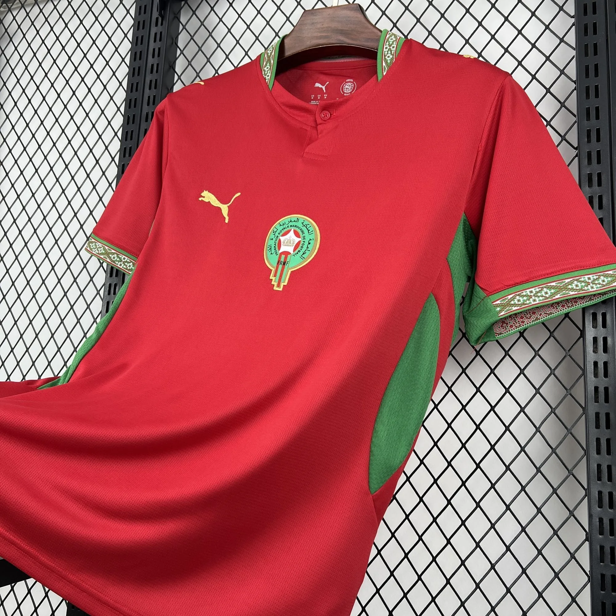 Vista en ángulo de la parte delantera de la camiseta roja, mostrando el cuello de botón con motivos verdes, el escudo de la federación en el pecho y el panel verde transpirable en el lateral.