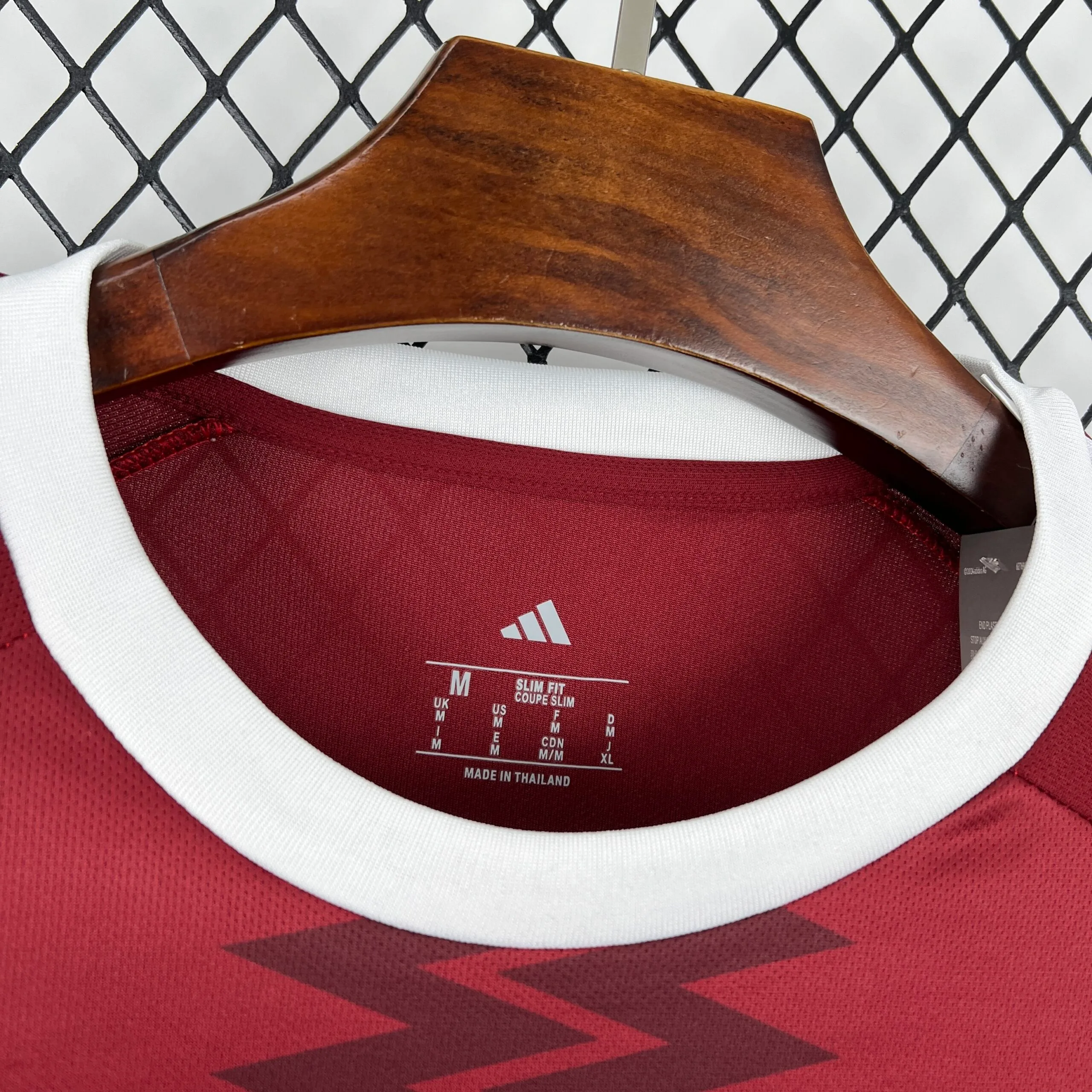 Primer plano del cuello redondo blanco de la camiseta de la selección de Qatar, mostrando en el interior el logotipo de la marca, las especificaciones de "SLIM FIT / COUPE SLIM", la talla "M" y la procedencia de fabricación sobre el tejido granate.