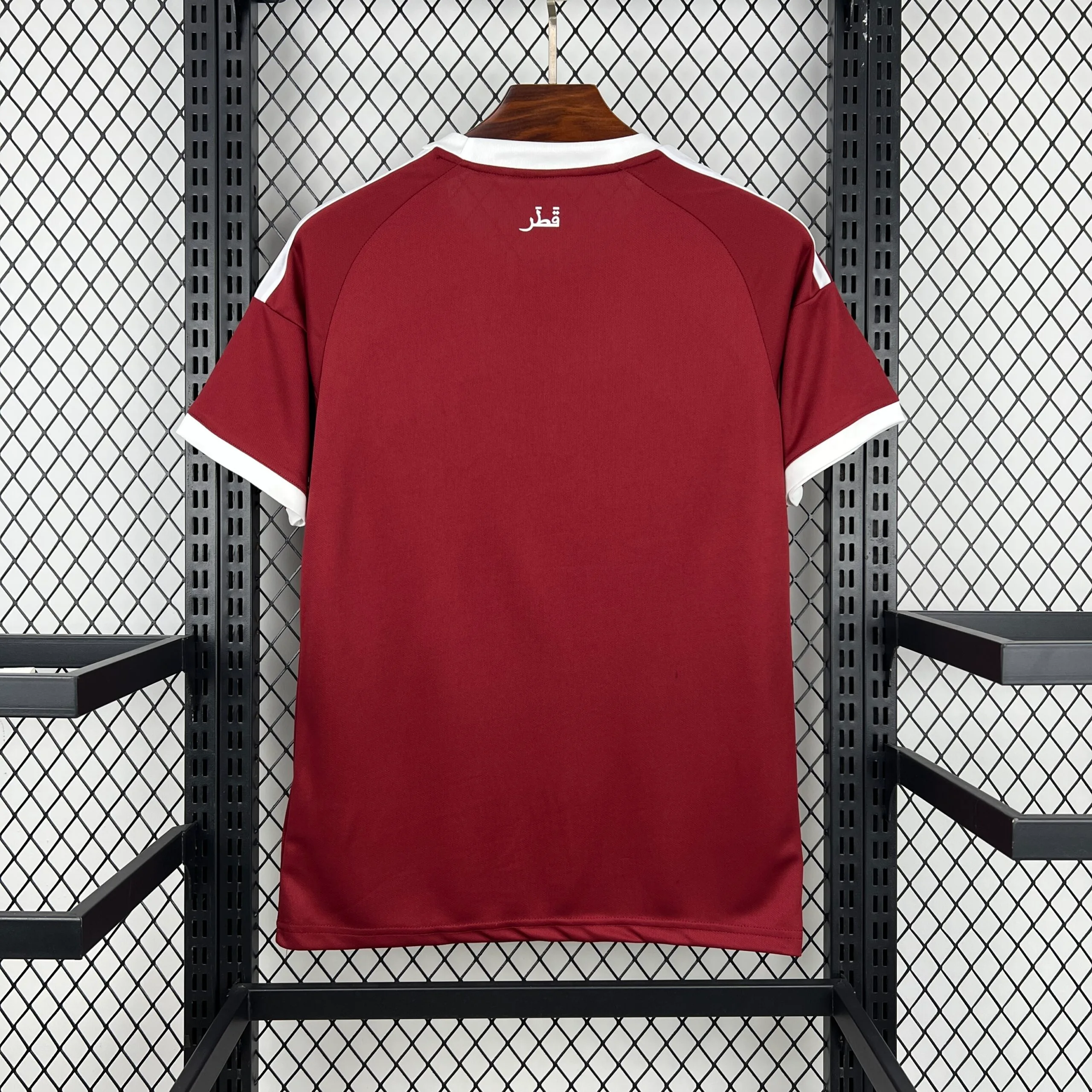 Parte posterior de la camiseta de la selección de Qatar en color granate liso, con cuello y bordes de las mangas en blanco, destacando el nombre del país escrito en caligrafía árabe ("قطر") en la zona de la nuca.