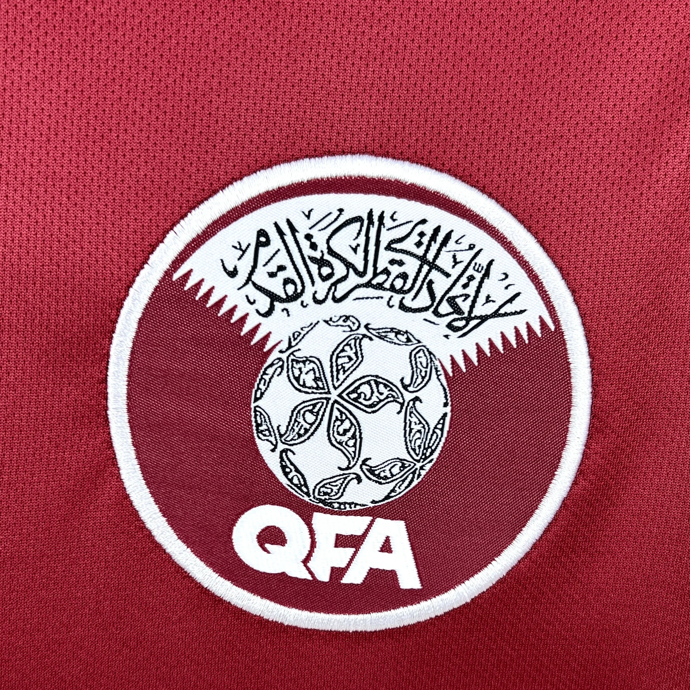 Primer plano del escudo circular de la Asociación de Fútbol de Qatar (QFA) bordado sobre el tejido granate. El emblema presenta un diseño en blanco y granate con un balón de fútbol decorado con patrones tradicionales, caligrafía árabe en la parte superior y las siglas "QFA" en la base.