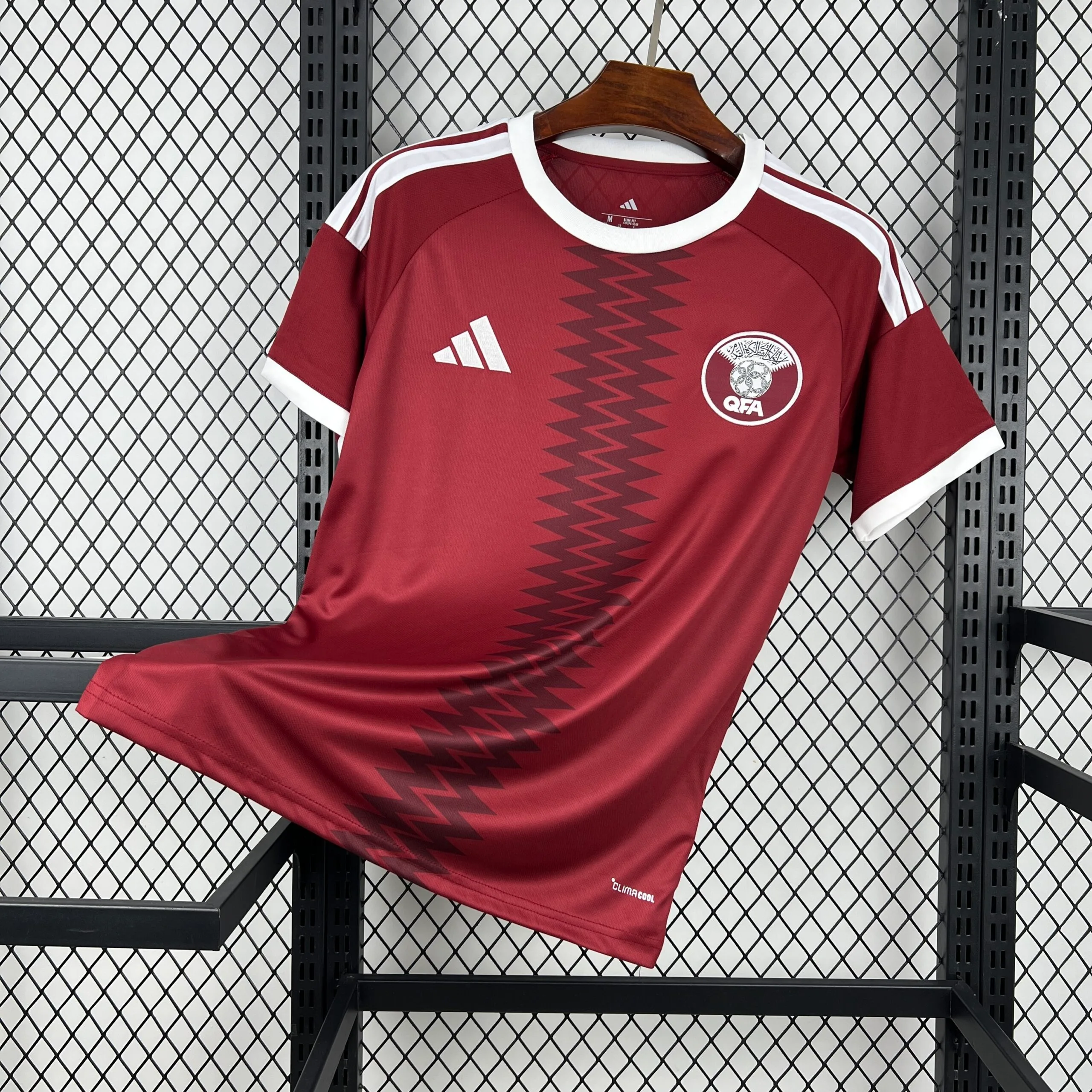 Camiseta de la selección de Qatar en color granate intenso con una franja vertical central de diseño en zigzag tonal. Presenta cuello redondo y bordes de las mangas en color blanco, con el logotipo de la marca y el escudo de la Asociación de Fútbol de Qatar (QFA) en el pecho.