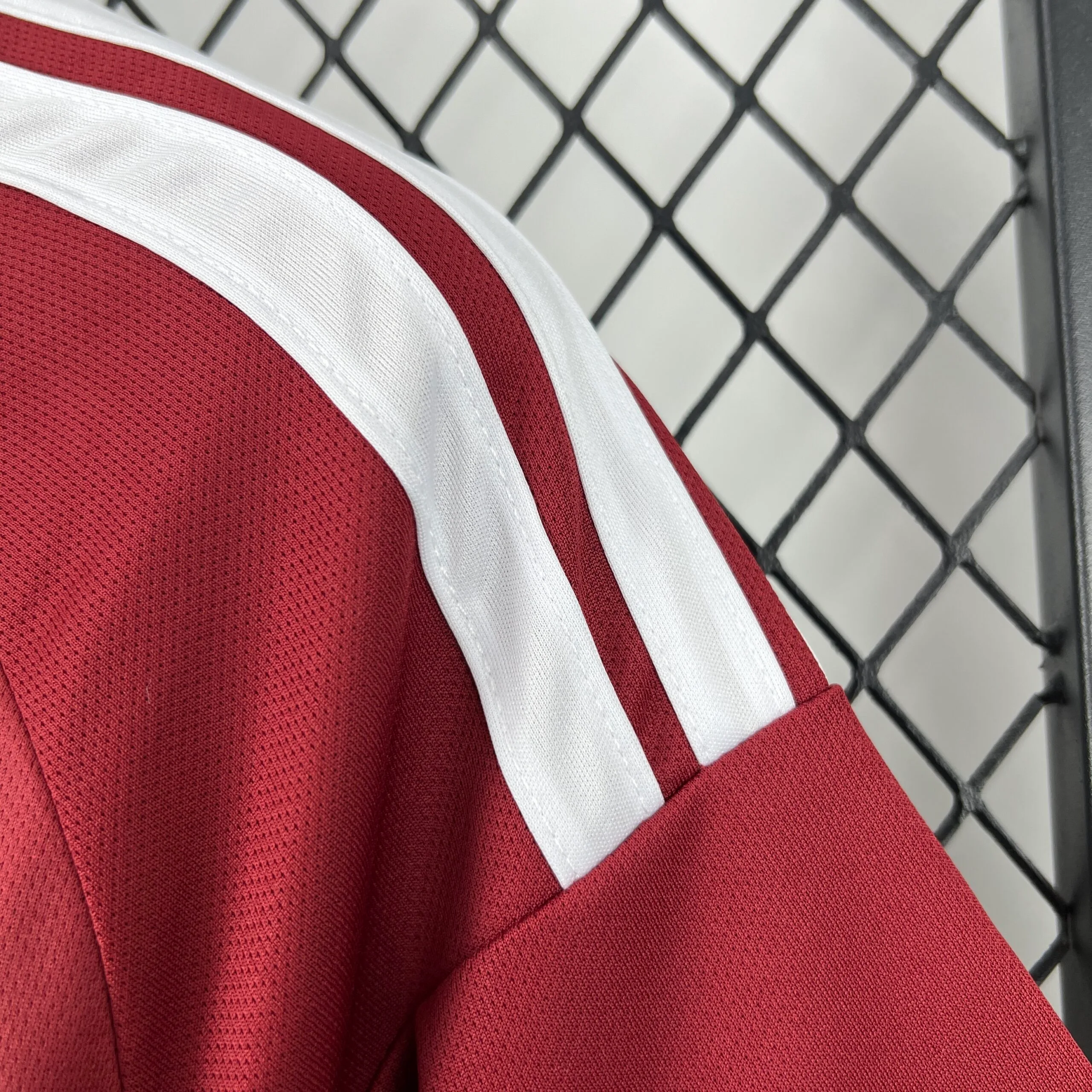 Primer plano de la parte superior de la manga y el hombro de la camiseta de la selección de Qatar, destacando el tejido granate y las tres franjas blancas paralelas que bajan desde el cuello.