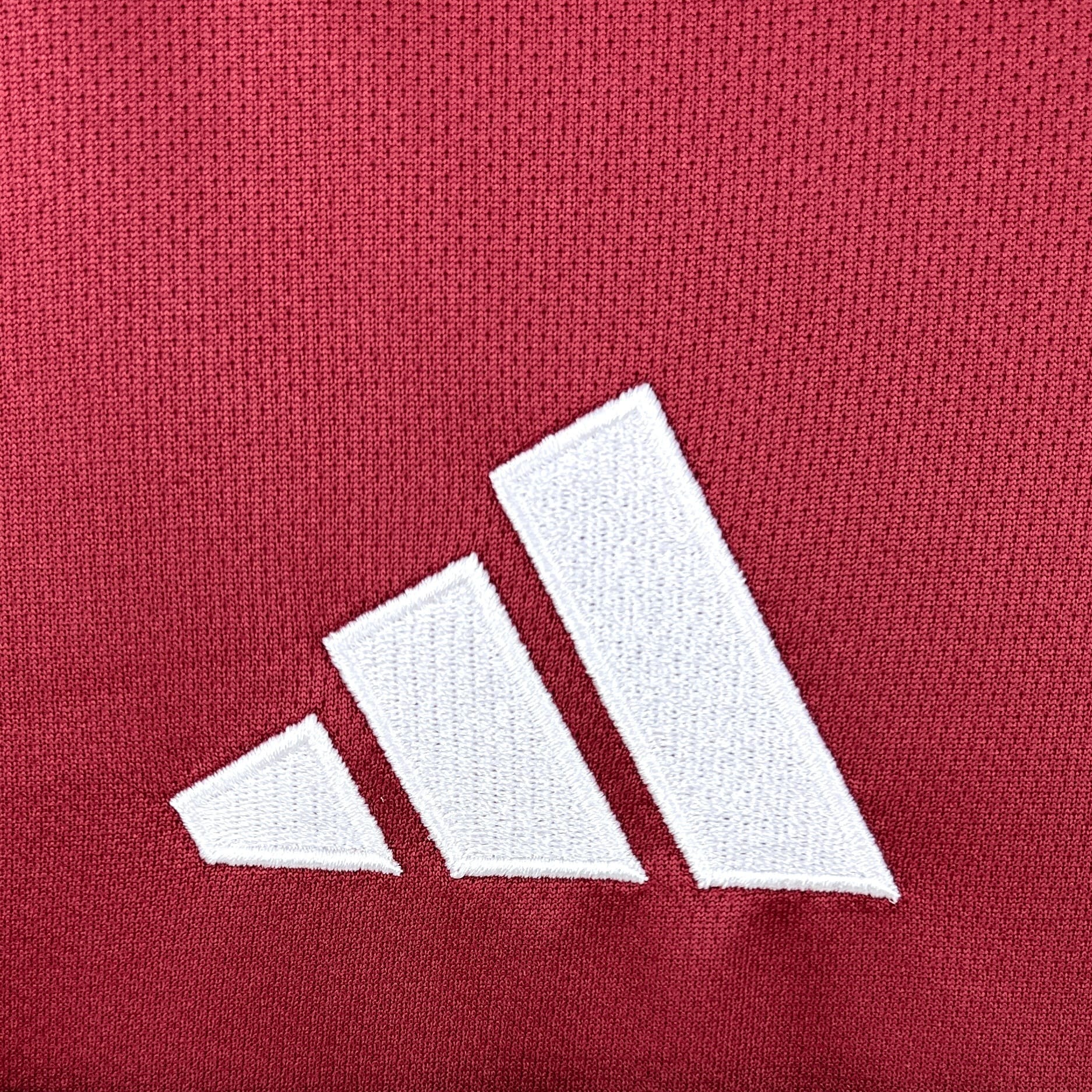 Primer plano de un logotipo compuesto por tres franjas diagonales blancas de tamaño ascendente, bordado con precisión sobre el tejido técnico de color granate de la camiseta de la selección de Qatar.