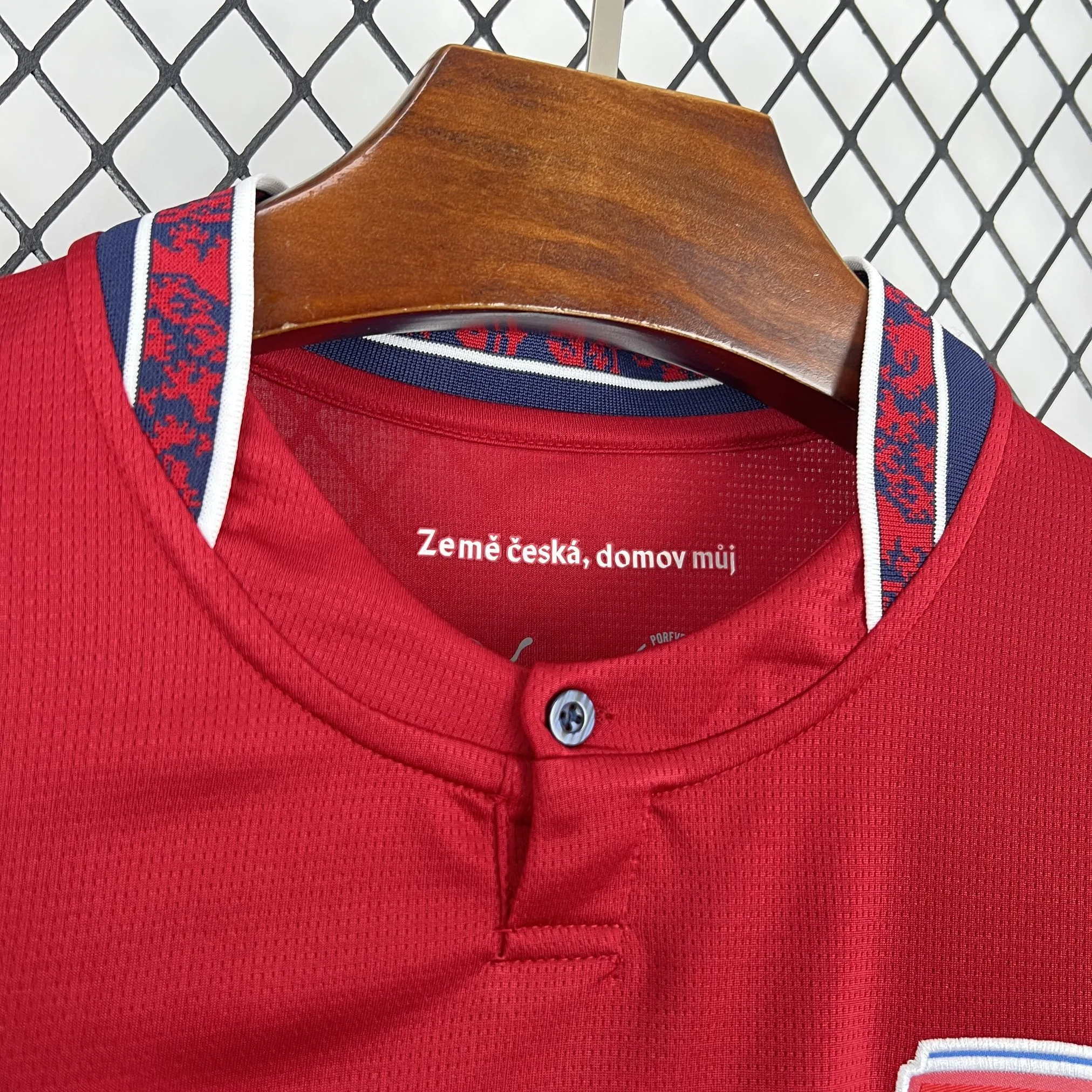 Detalle del cuello tipo polo con cierre de un botón y la inscripción "Země česká, domov můj" estampada en el interior de la nuca sobre el tejido técnico rojo.