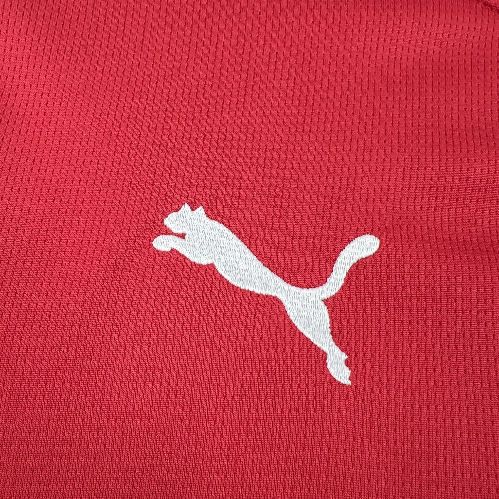 Detalle del logotipo de la marca bordado en hilo blanco sobre la textura microperforada del tejido rojo de la camiseta checa.
