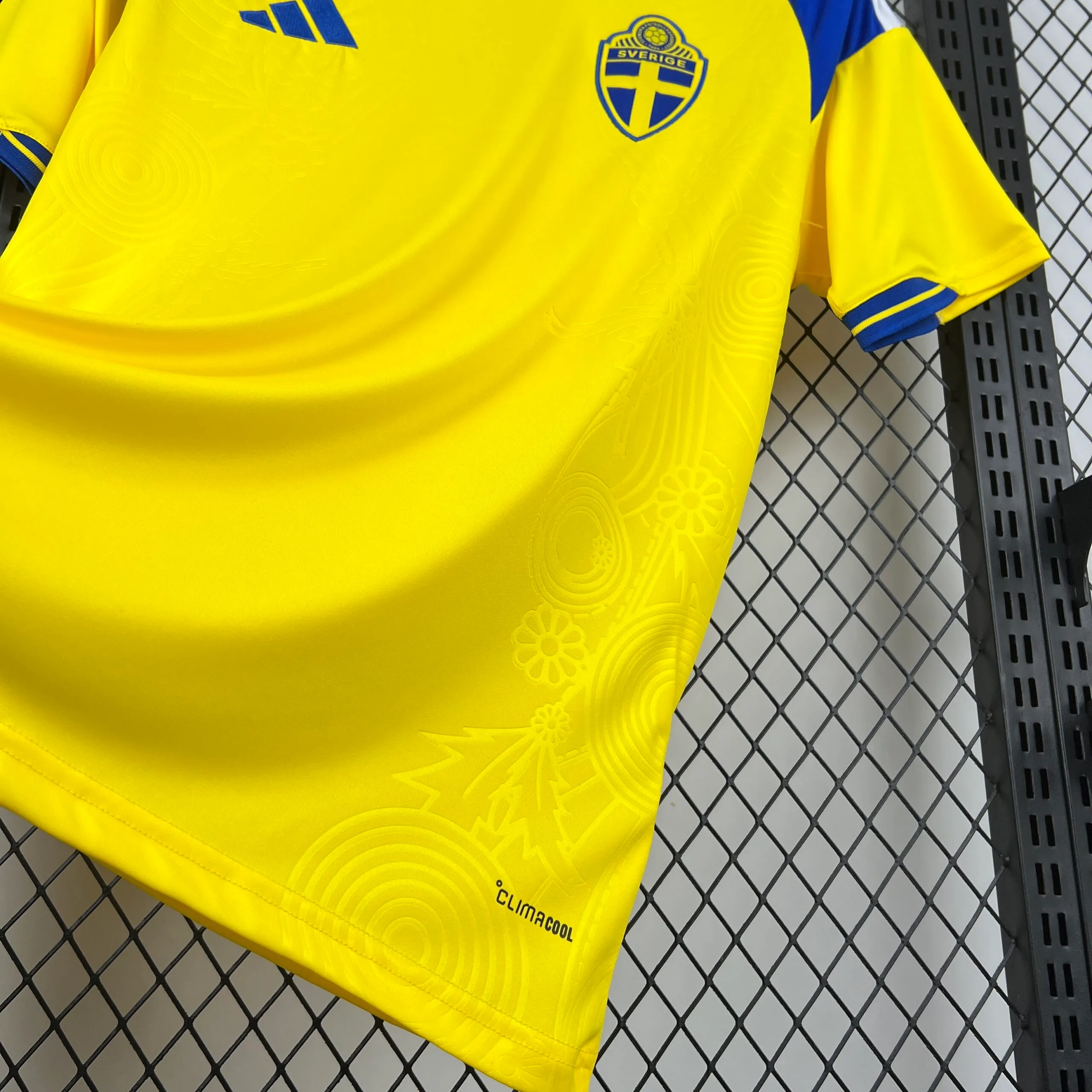 Vista en ángulo de la parte inferior de la camiseta amarilla, resaltando la profundidad del patrón tonal grabado con flores y círculos. En la esquina inferior derecha se aprecia el sello de la tecnología "CLIMACOOL" en letras negras sobre el tejido técnico.