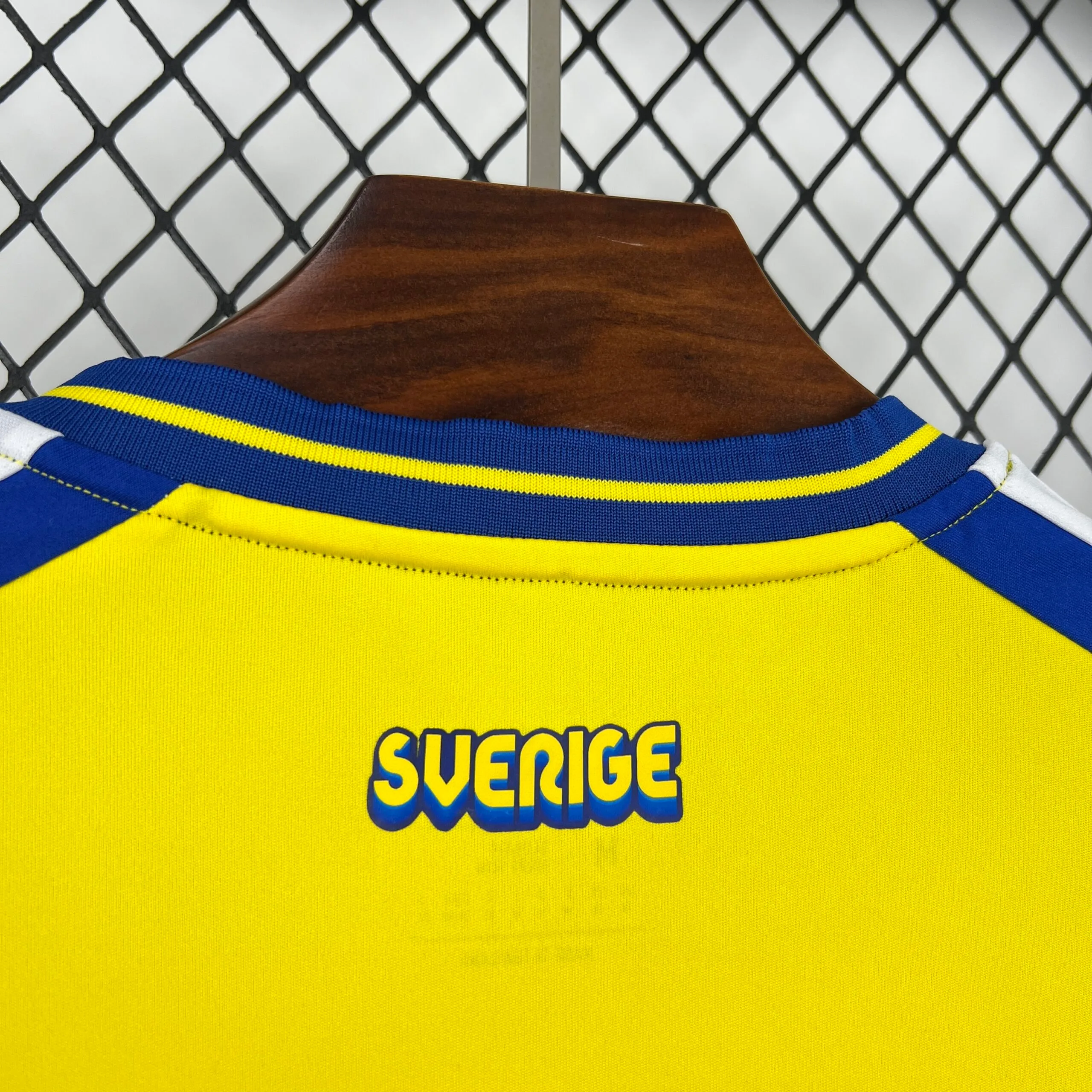 Primer plano de la parte posterior del cuello de la camiseta, resaltando la inscripción "SVERIGE" estampada en letras amarillas con un marcado efecto de sombra en azul real. El detalle se sitúa justo debajo del ribete azul del cuello y sobre el tejido amarillo liso de la espalda.