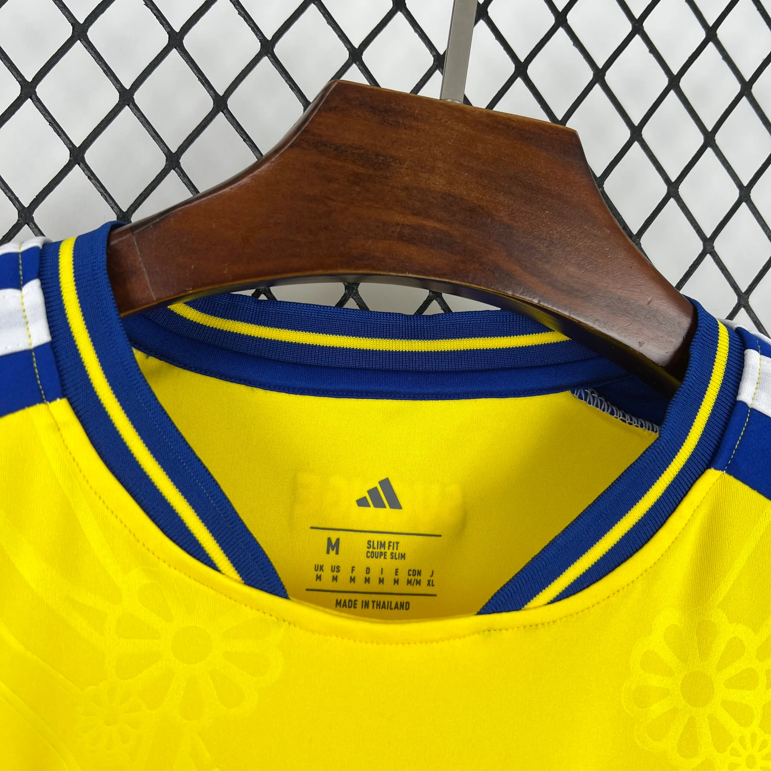 Primer plano del interior del cuello azul con ribetes amarillos de la camiseta de Suecia. En el tejido amarillo con patrón floral se observa el logotipo de la marca, la talla "M", el corte "SLIM FIT" y el sello de fabricación "MADE IN THAILAND" impresos en color negro.