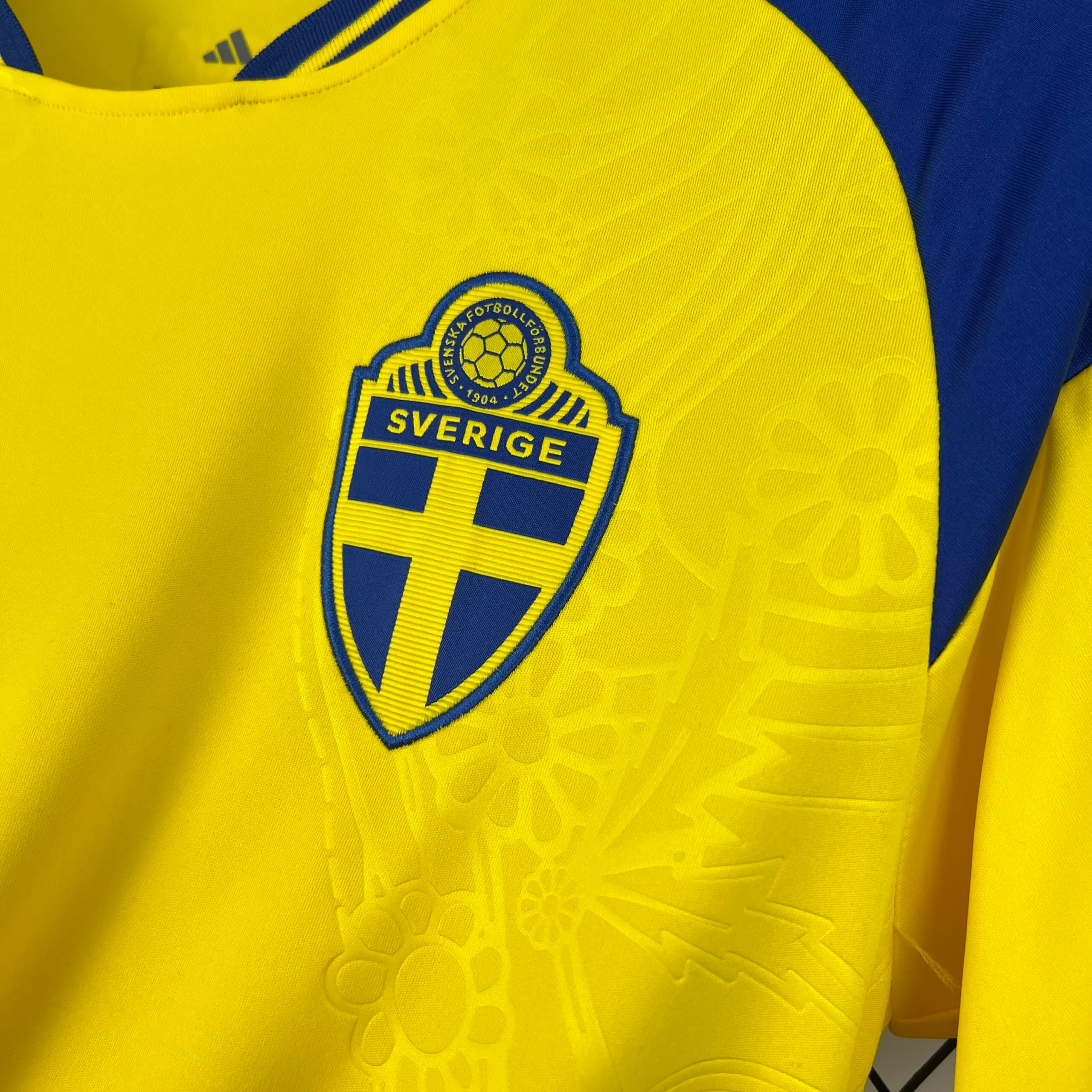Primer plano del escudo de la federación sueca bordado en el pecho. Presenta una forma de escudo azul con la bandera de Suecia (cruz amarilla), coronado por un balón de fútbol antiguo y la palabra "SVERIGE" en la parte superior. El escudo está rodeado por el patrón tonal de flores y círculos grabados en el tejido amarillo.
