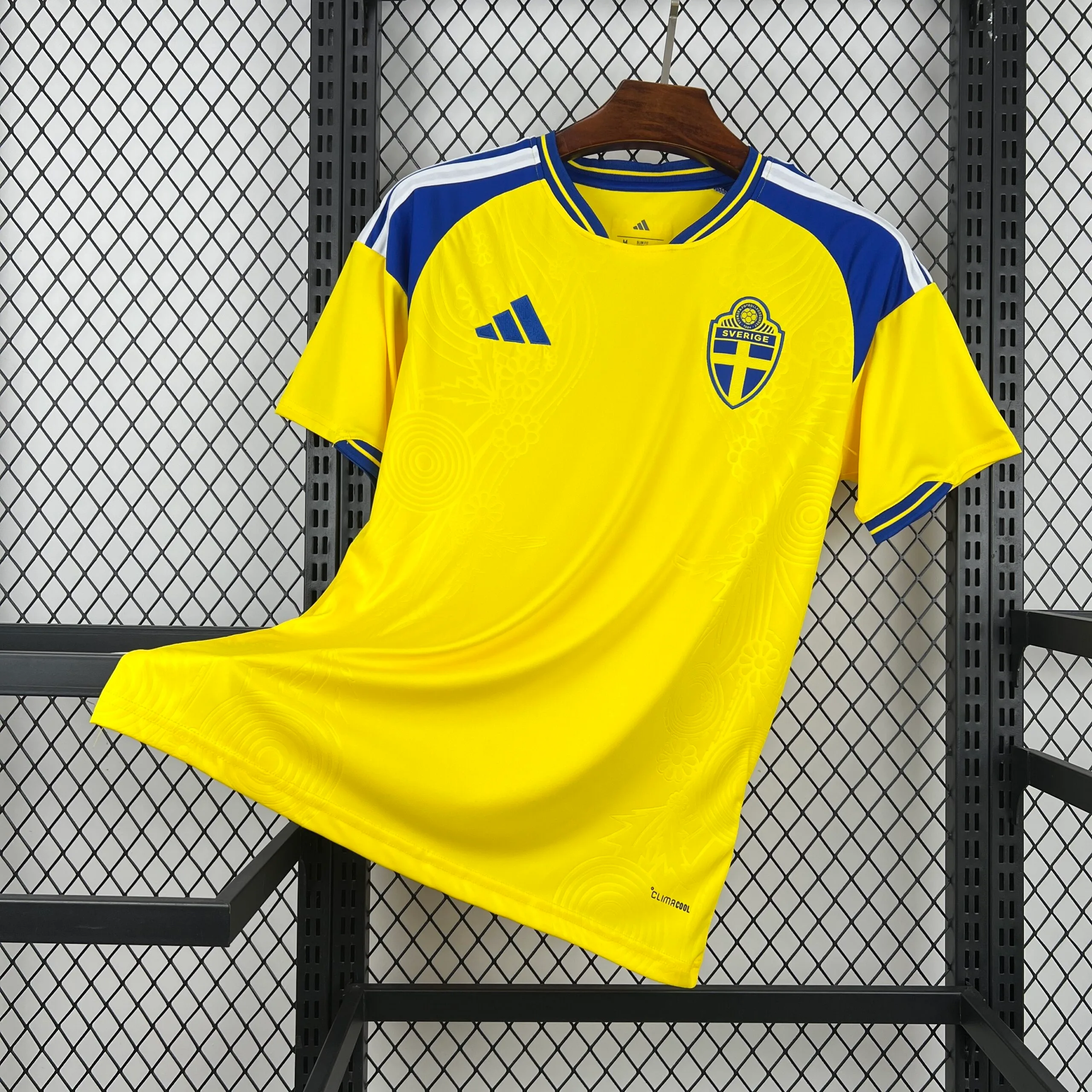 Camiseta en color amarillo vibrante con un patrón tonal de círculos y flores grabados en el tejido. Presenta detalles en azul real en los hombros, el cuello redondo y los puños, además de las tres franjas blancas sobre los hombros. En el pecho se encuentran el logotipo de la marca en azul y el escudo de la selección sueca con la inscripción "SVERIGE".