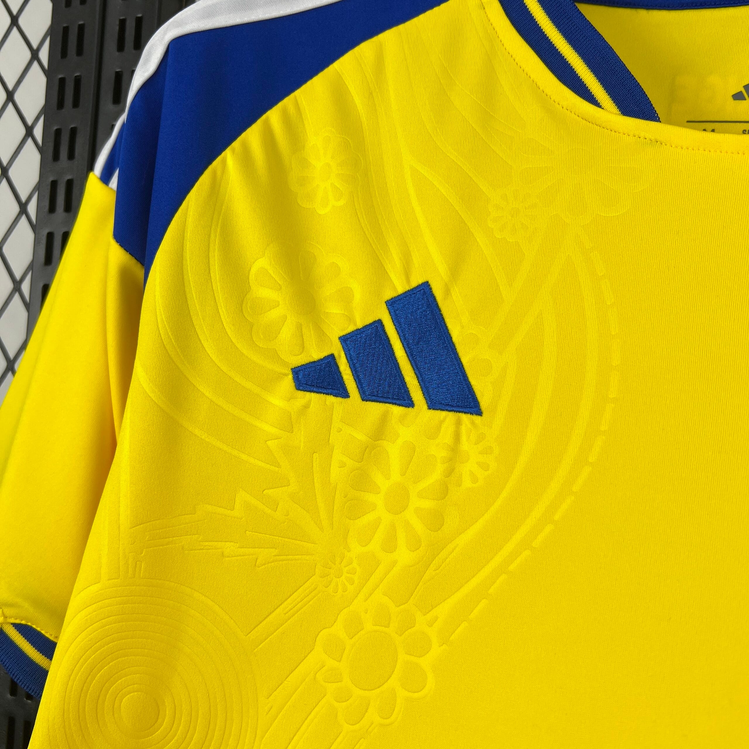 Primer plano del logotipo de la marca deportiva bordado en azul real sobre el tejido amarillo. La imagen destaca el patrón tonal de flores y círculos grabados en relieve que decora toda la camiseta, así como el panel azul del hombro en la parte superior.