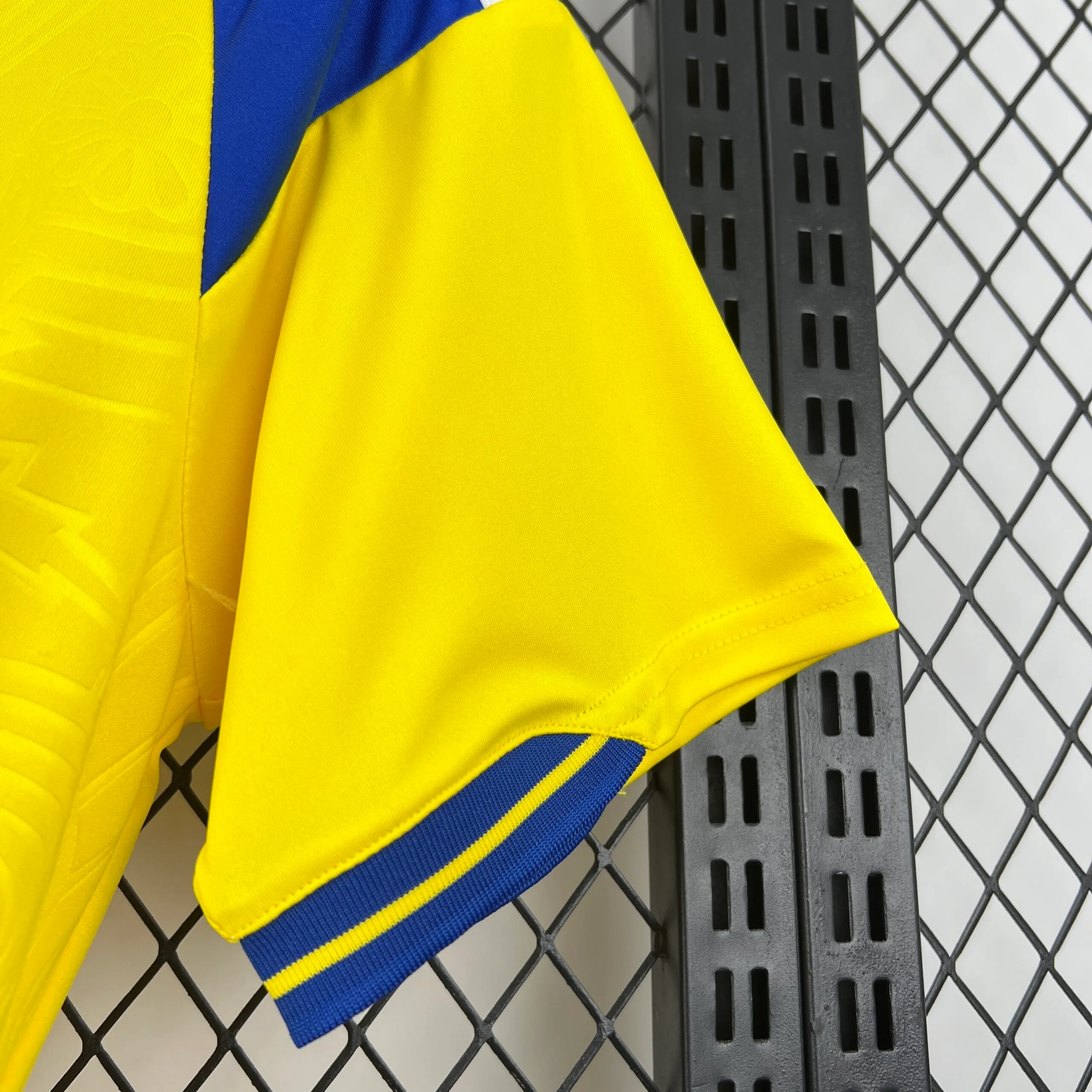 Primer plano de la manga derecha en color amarillo, destacando el acabado del puño en tejido elástico azul con una franja amarilla central. En el fondo se aprecia el panel azul del hombro y la estructura de rejilla metálica negra donde está colgada la prenda.
