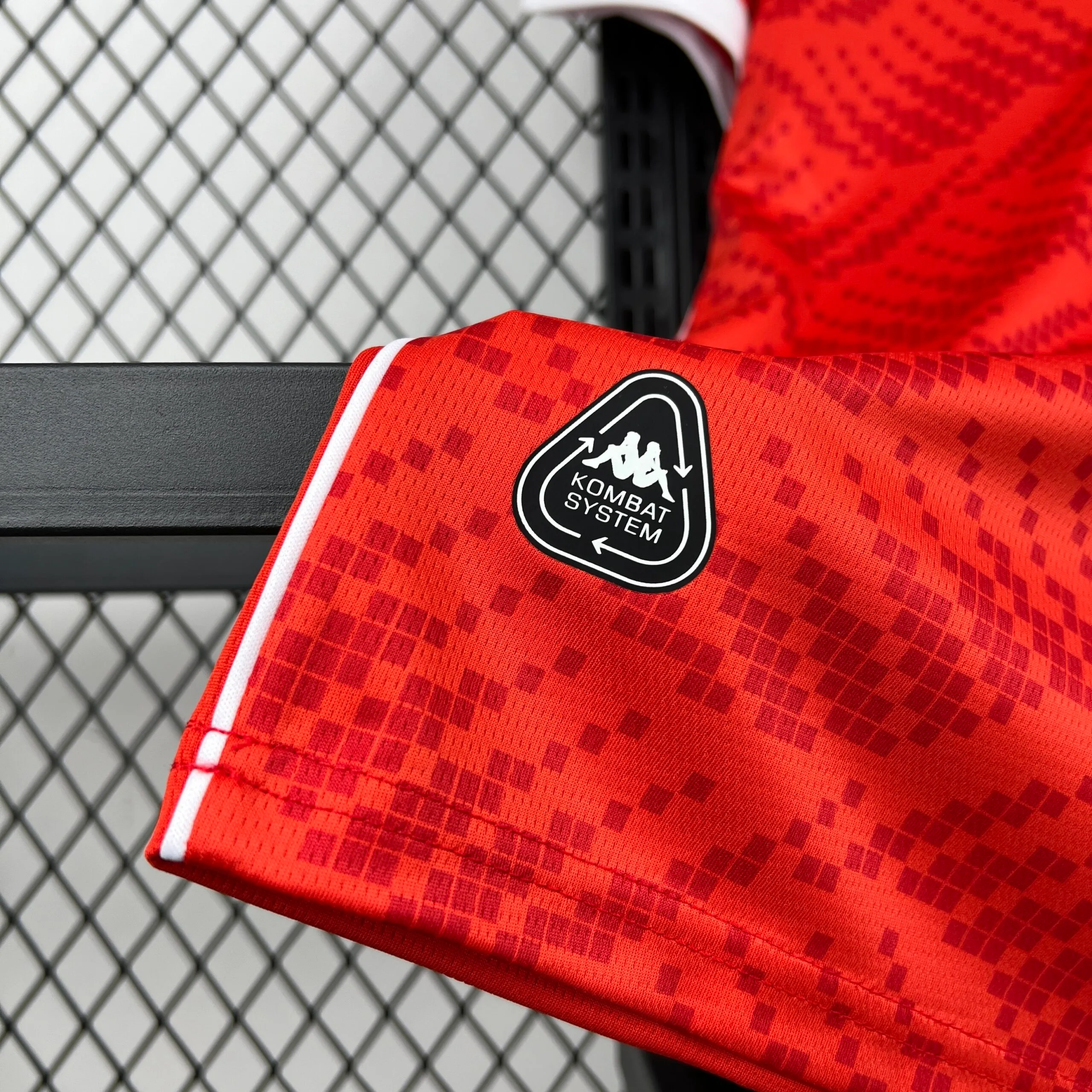 Primer plano de la parte inferior de la camiseta de la selección de Túnez, que muestra el tejido rojo con patrón de diamantes, un fino ribete blanco lateral y el sello de tecnología "KOMBAT SYSTEM" en un parche termosellado de color negro.
