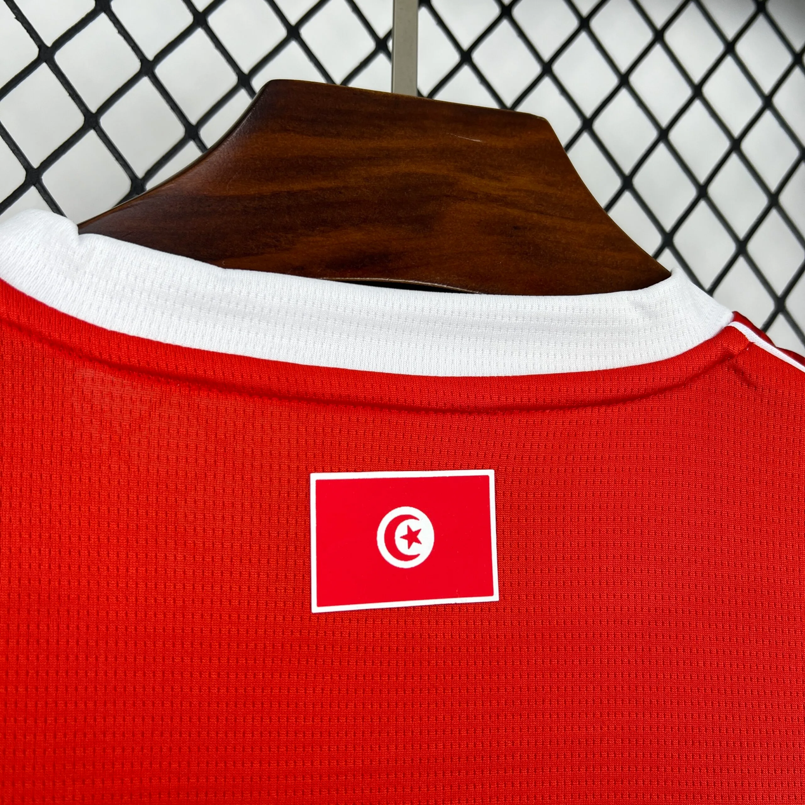 Primer plano de la parte posterior del cuello blanco de la camiseta de la selección de Túnez, destacando una pequeña bandera nacional rectangular estampada sobre el tejido técnico rojo.