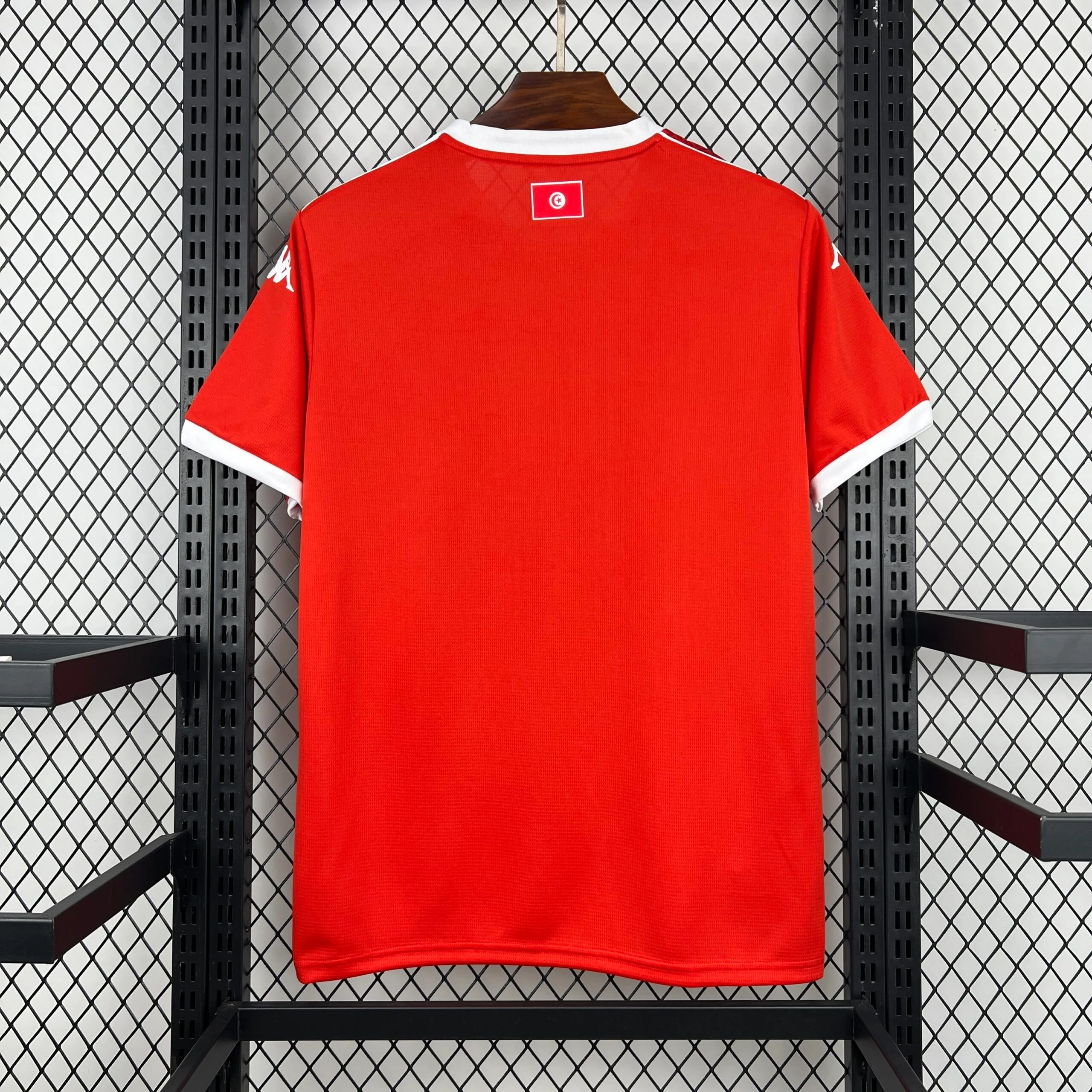 Parte posterior de la camiseta de la selección de Túnez en color rojo liso, destacando el cuello redondo y los bordes de las mangas en color blanco, junto con una pequeña bandera nacional en la zona de la nuca.
