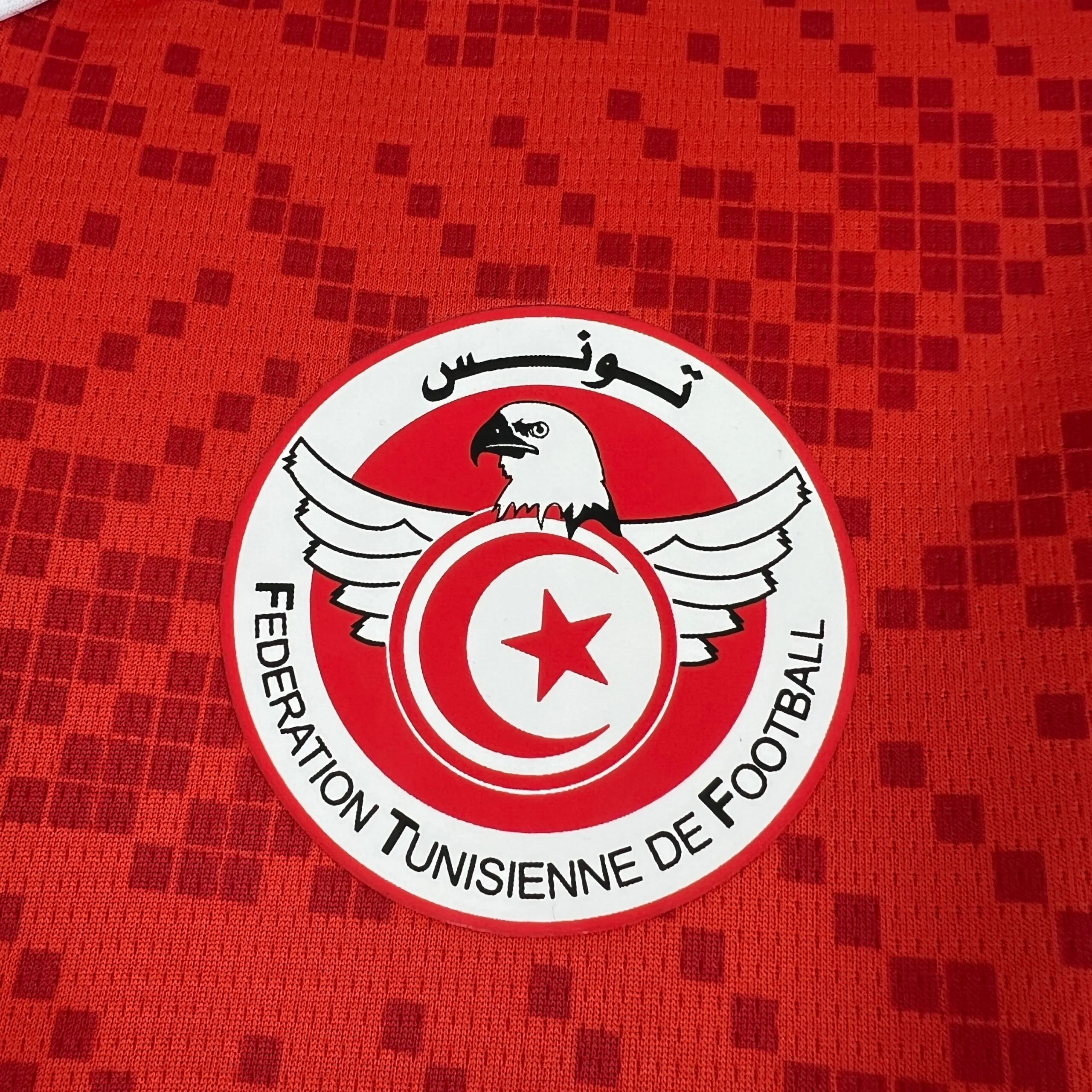 Primer plano del escudo circular de la Federación Tunecina de Fútbol sobre el tejido rojo con patrón de diamantes. El emblema muestra un águila blanca con las alas extendidas protegiendo un círculo rojo con la media luna y la estrella, rodeado por el nombre de la federación en francés y árabe.