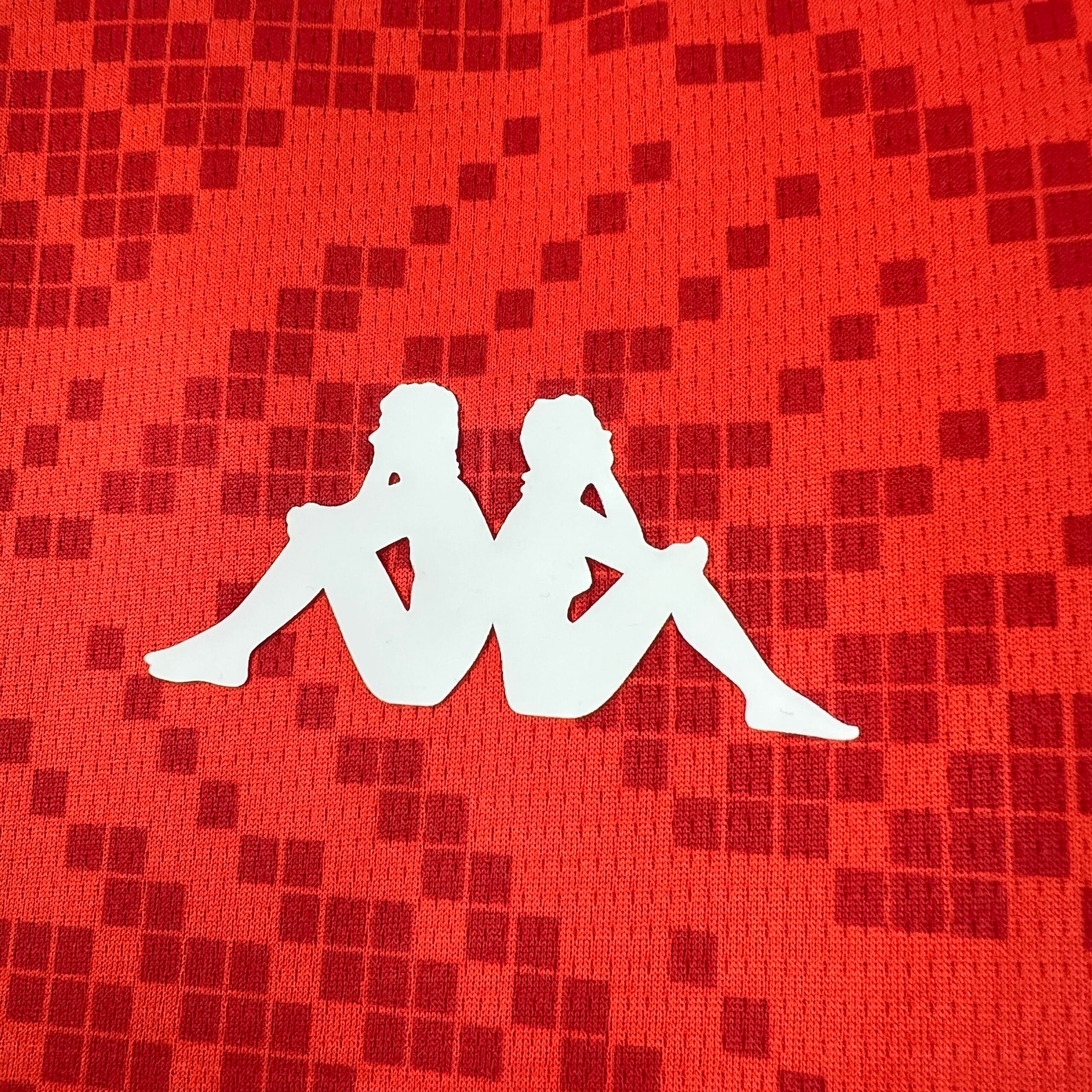 Primer plano de un logotipo blanco que muestra las siluetas de dos figuras sentadas espalda con espalda, estampado sobre el tejido rojo con patrón de diamantes de la camiseta de la selección de Túnez.