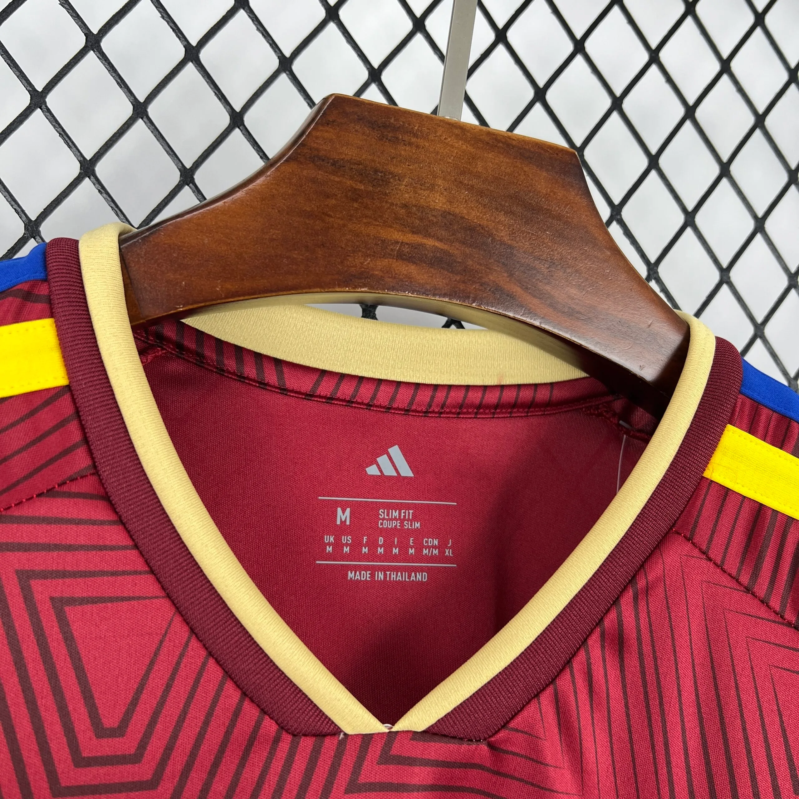 Primer plano del cuello en "V" de la camiseta de la selección de Venezuela, destacando el ribete exterior en color crema y el interior en vino tinto. En la parte interna se aprecia el logotipo de la marca, la talla "M", las especificaciones "SLIM FIT / COUPE SLIM" y su origen de fabricación sobre el tejido con patrón geométrico.