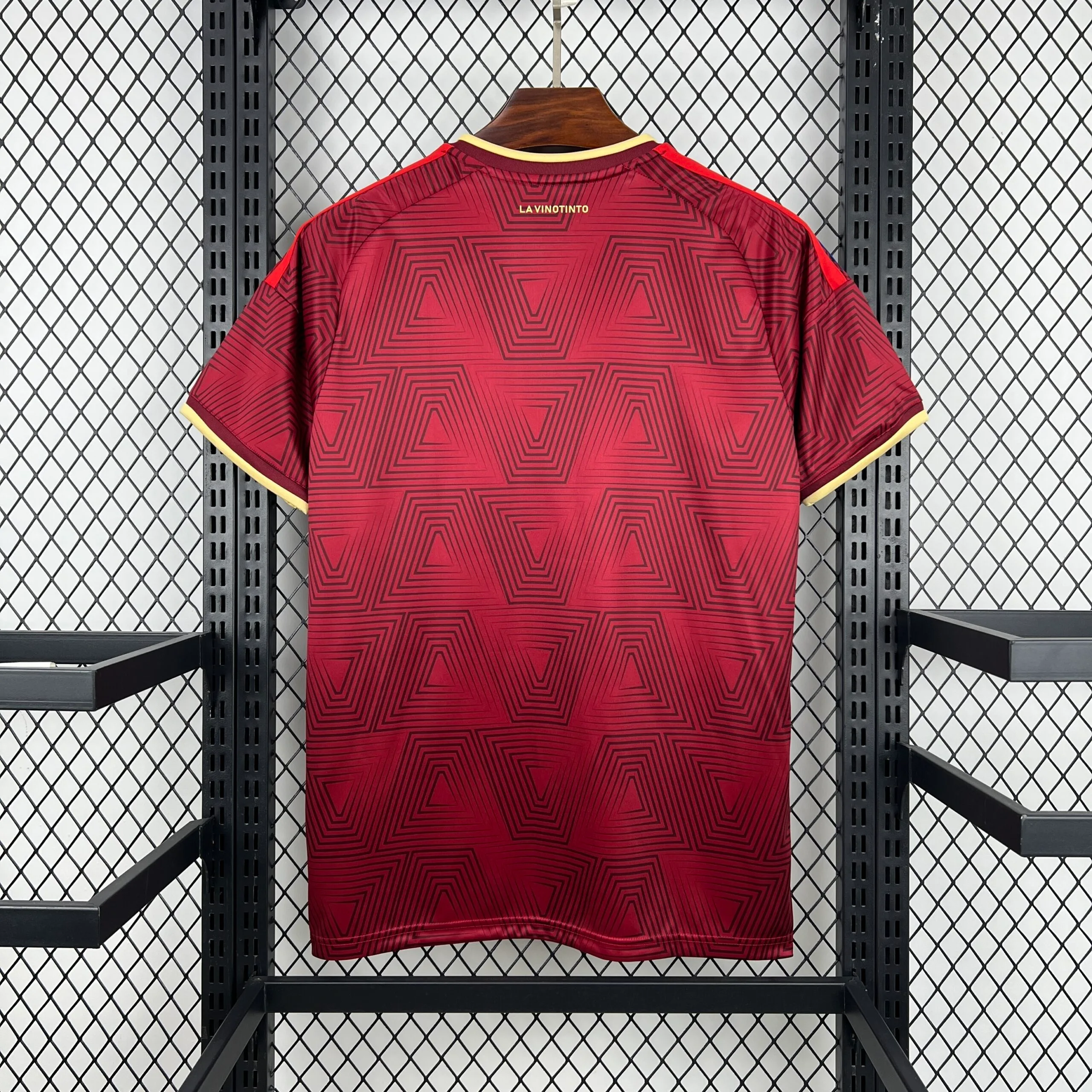 Parte posterior de la camiseta de la selección de Venezuela en color vino tinto, donde se aprecia la continuidad del patrón de cuadros concéntricos. En la zona de la nuca, destaca la frase "LA VINOTINTO" estampada en letras doradas sobre el borde del cuello crema.