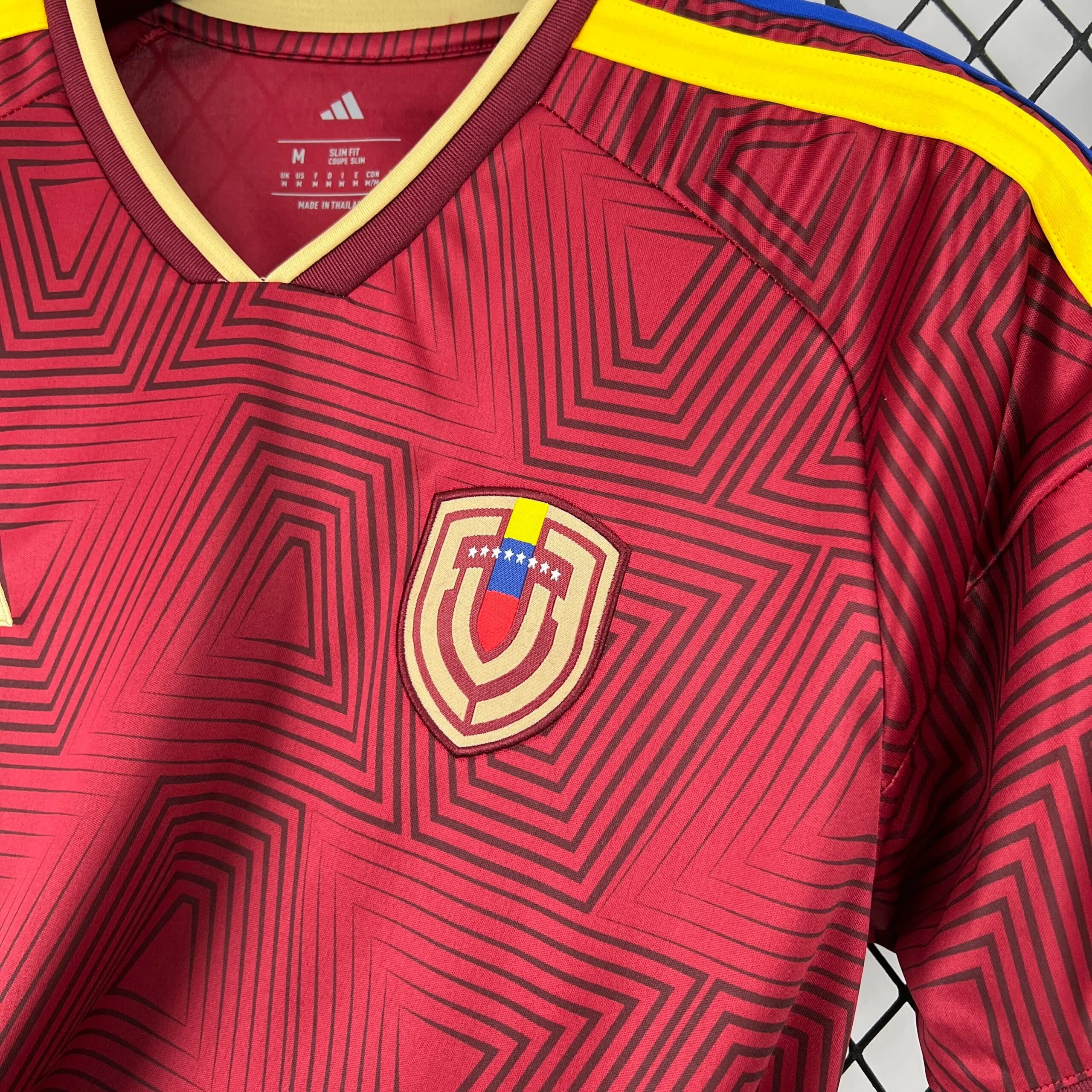 Primer plano del escudo de la Federación Venezolana de Fútbol sobre el tejido vino tinto con patrón de cuadros. El emblema, con borde crema, presenta las siglas "FVF" estilizadas e integra en su parte superior la bandera nacional con sus ocho estrellas blancas sobre la franja azul.