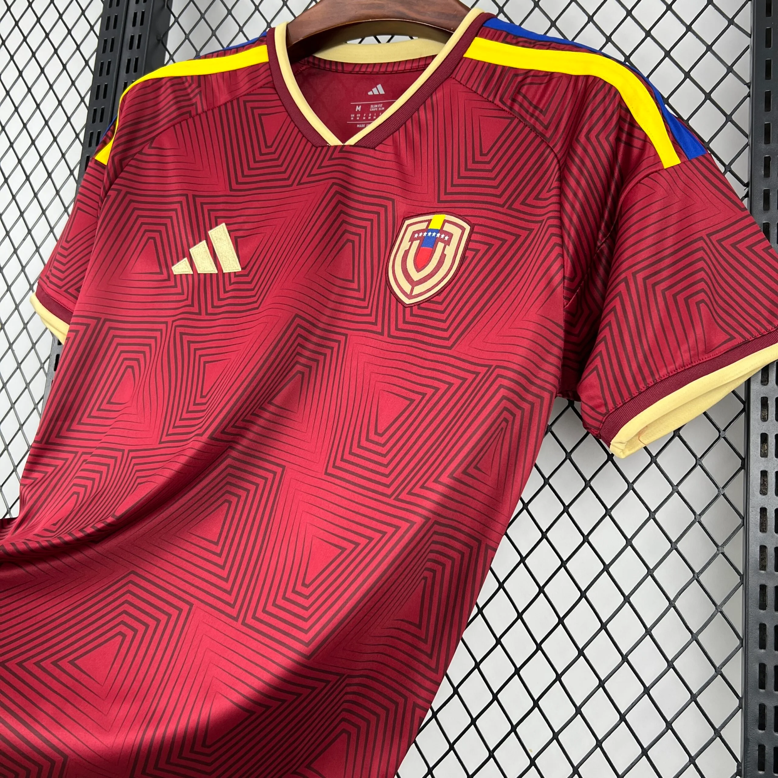 Perspectiva de la camiseta de la selección de Venezuela en color vino tinto, resaltando el patrón de cuadros concéntricos, el escudo de la federación en el pecho y el diseño de las franjas amarillas y azules sobre los hombros que evocan la bandera nacional.