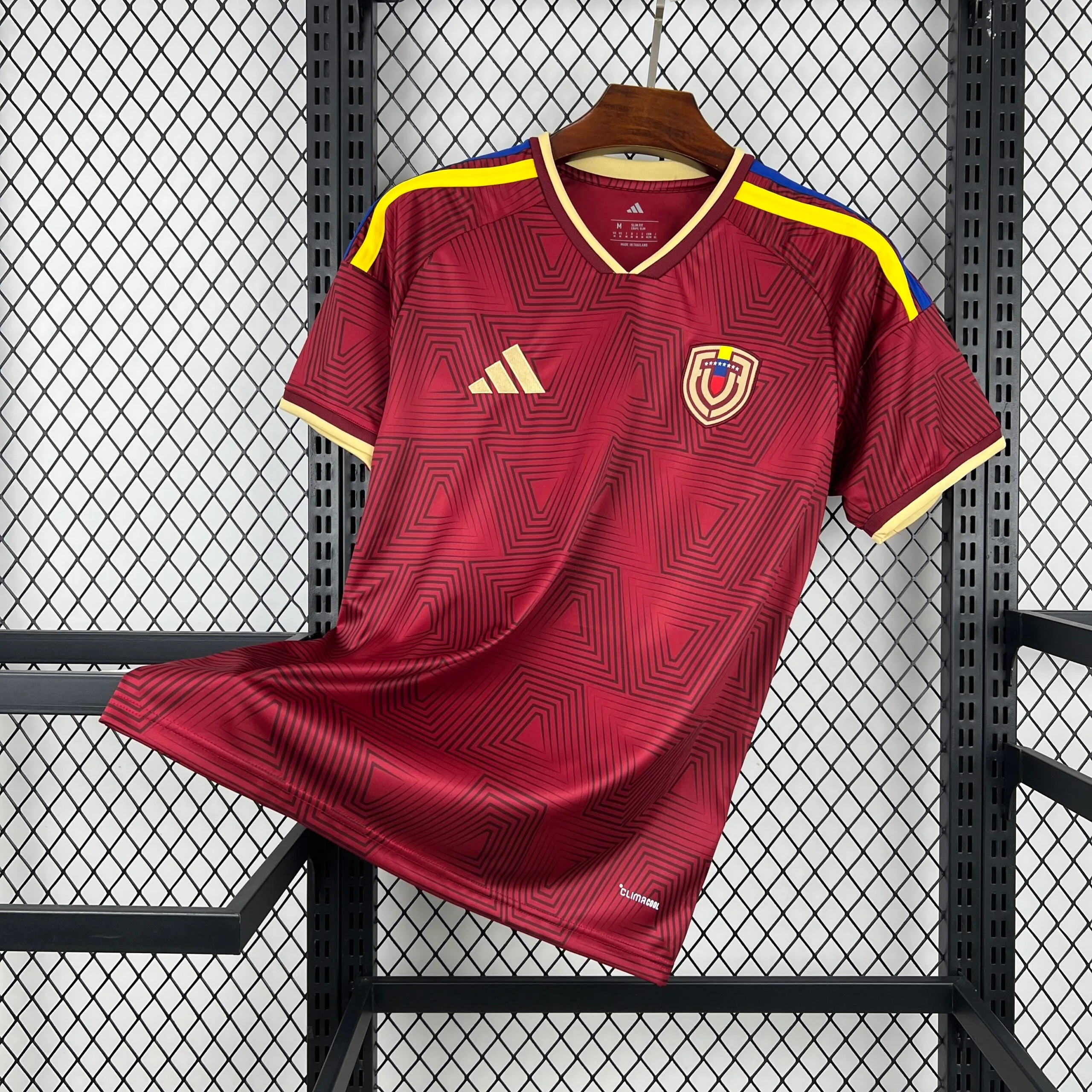 Camiseta de la selección de Venezuela en color vino tinto con un patrón gráfico de cuadrados concéntricos en degradado por toda la prenda. Presenta cuello en "V" con ribete crema, las tres franjas de los hombros en color amarillo y azul, el logotipo de la marca en dorado y el escudo de la federación venezolana en el pecho.