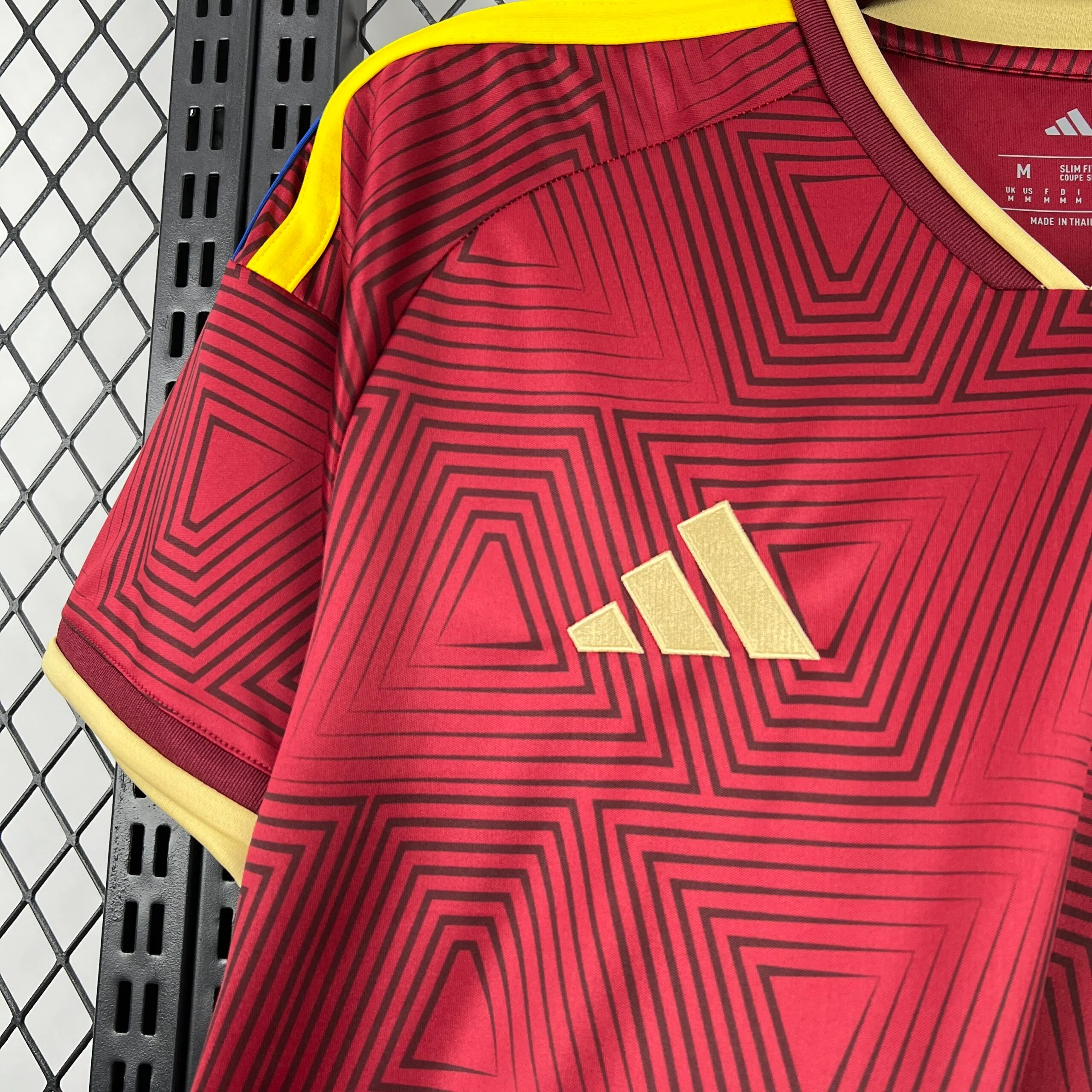 Primer plano del logotipo de la marca en color dorado, aplicado sobre el panel frontal de la camiseta de la selección de Venezuela, destacando el patrón de líneas geométricas en color vino tinto y las franjas amarillas del hombro.