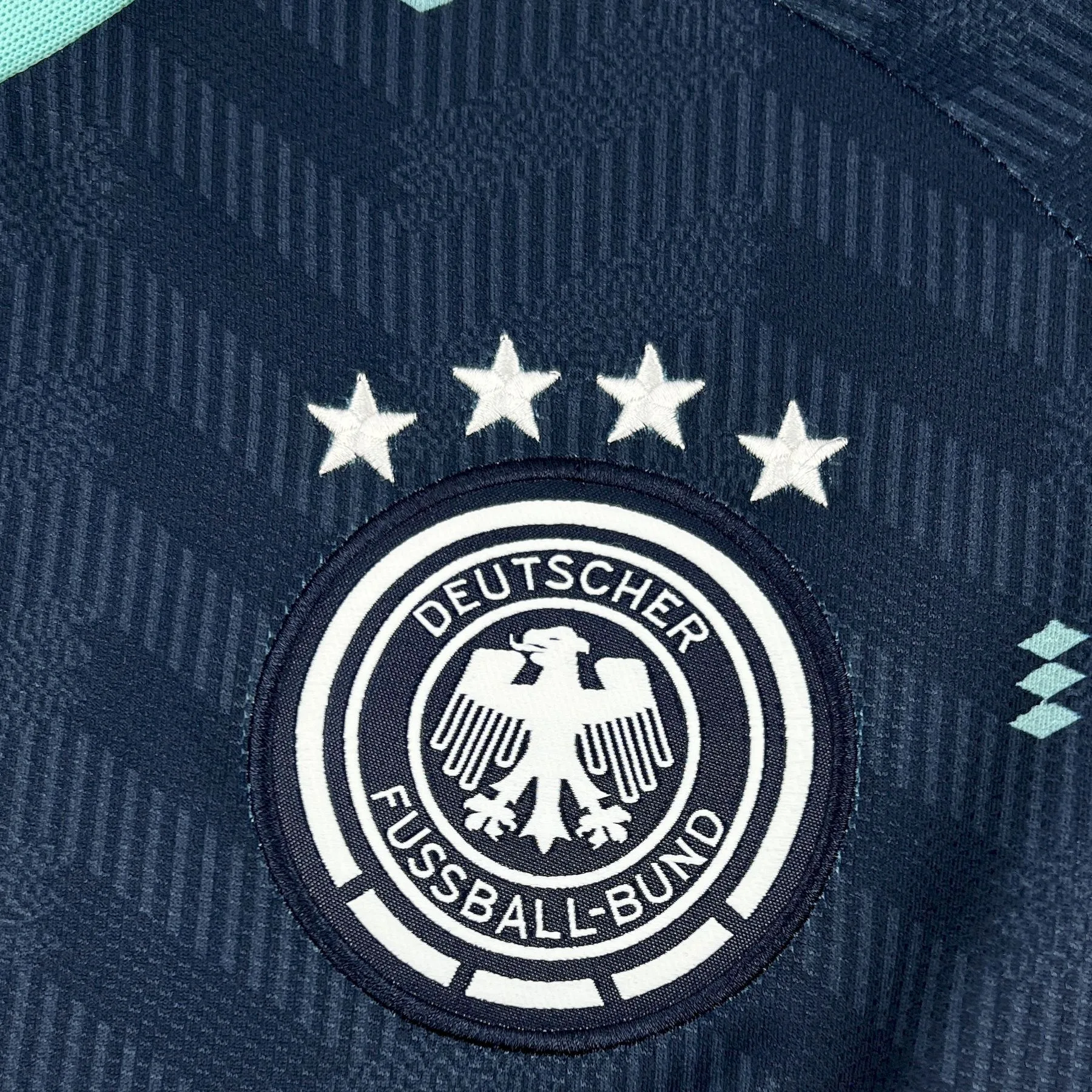 Detalle del escudo de la selección de Alemania bordado en blanco sobre el fondo rosa vibrante de la equipación.