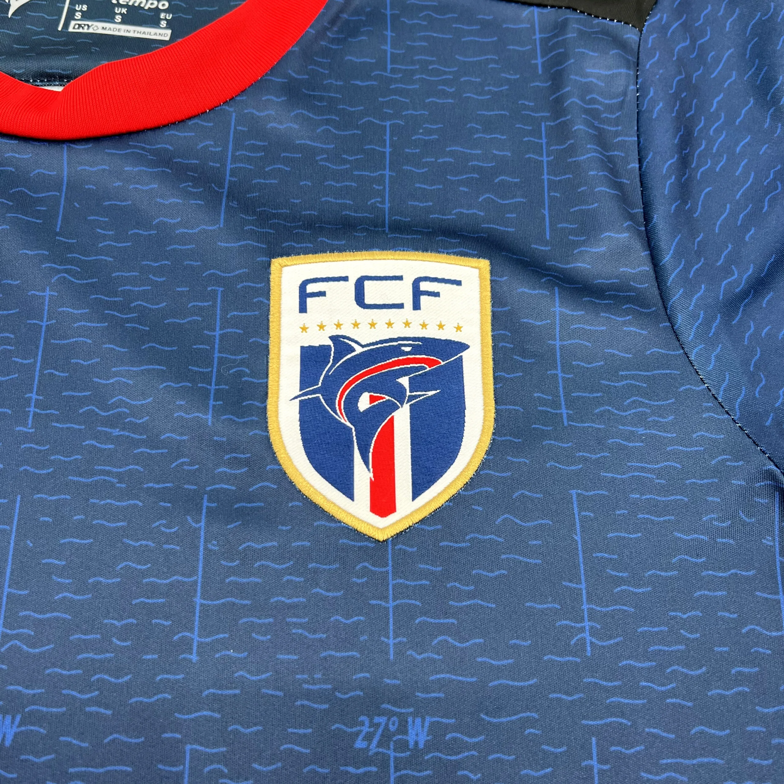 Primer plano del escudo de la Federación Caboverdiana de Fútbol (FCF) con el tiburón azul sobre fondo marino.