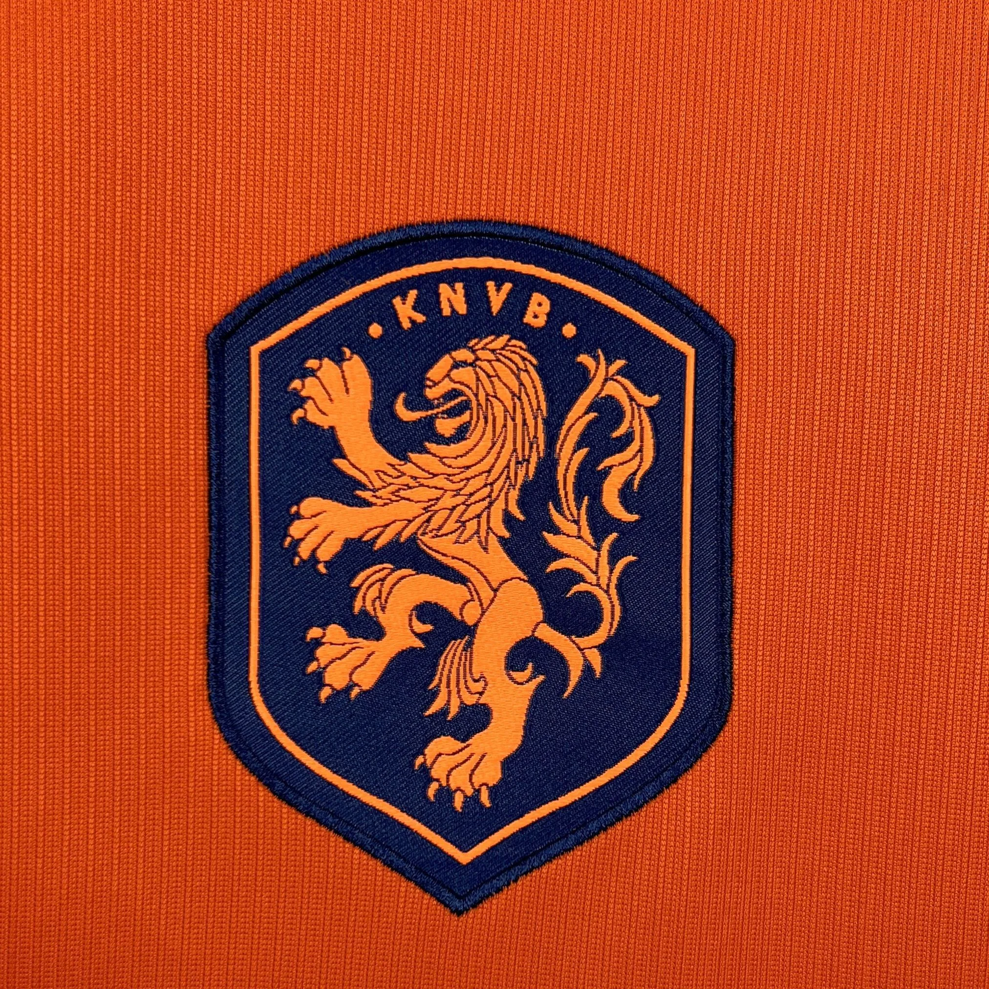 Primer plano del escudo del león de la KNVB bordado en negro sobre el tejido naranja de la camiseta.