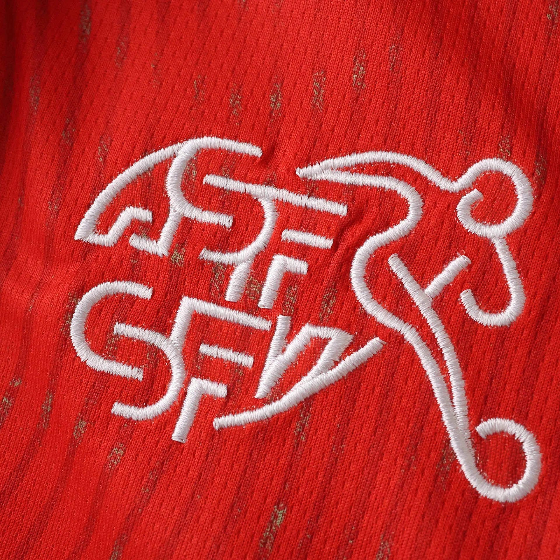Detalle del logotipo de la Asociación Suiza de Fútbol bordado en blanco con líneas dinámicas sobre fondo rojo.