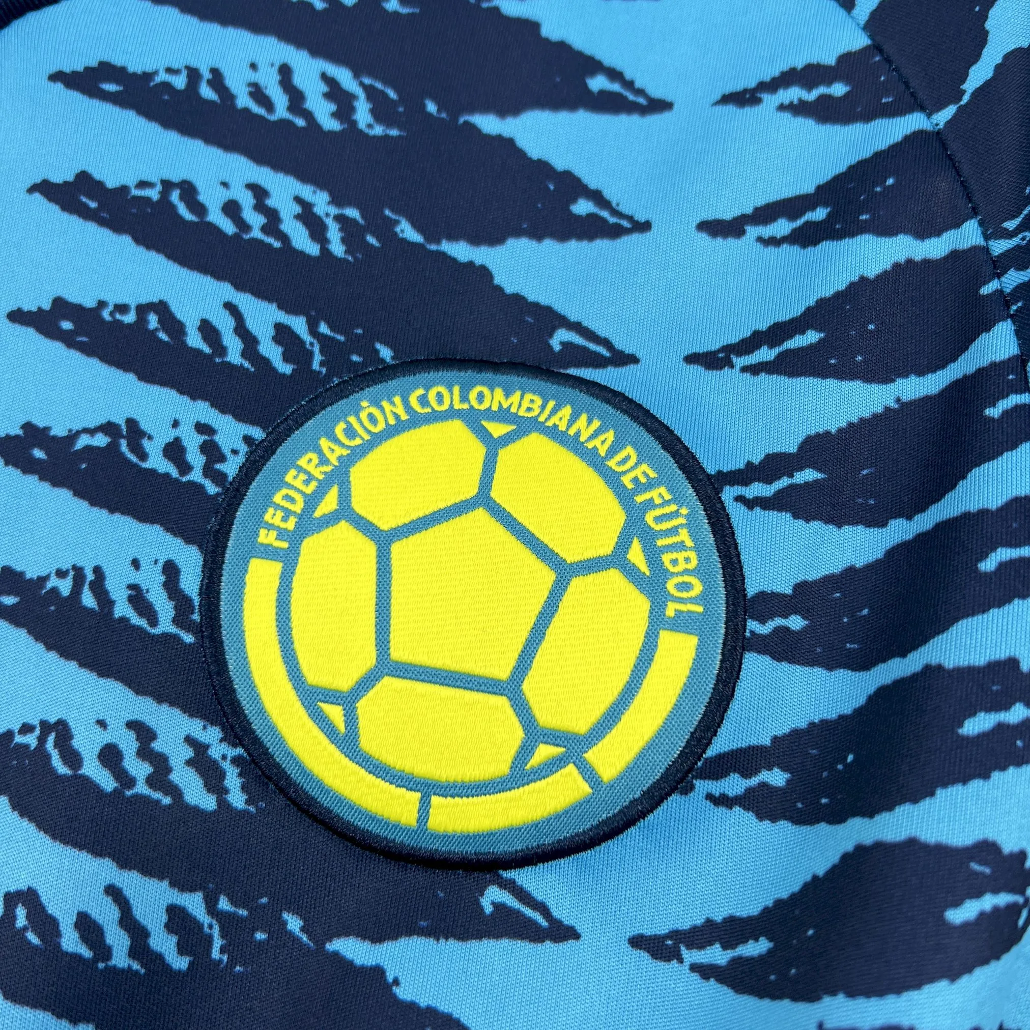 Primer plano del escudo de la Federación Colombiana de Fútbol bordado en tonos naranja y negro sobre el tejido técnico.