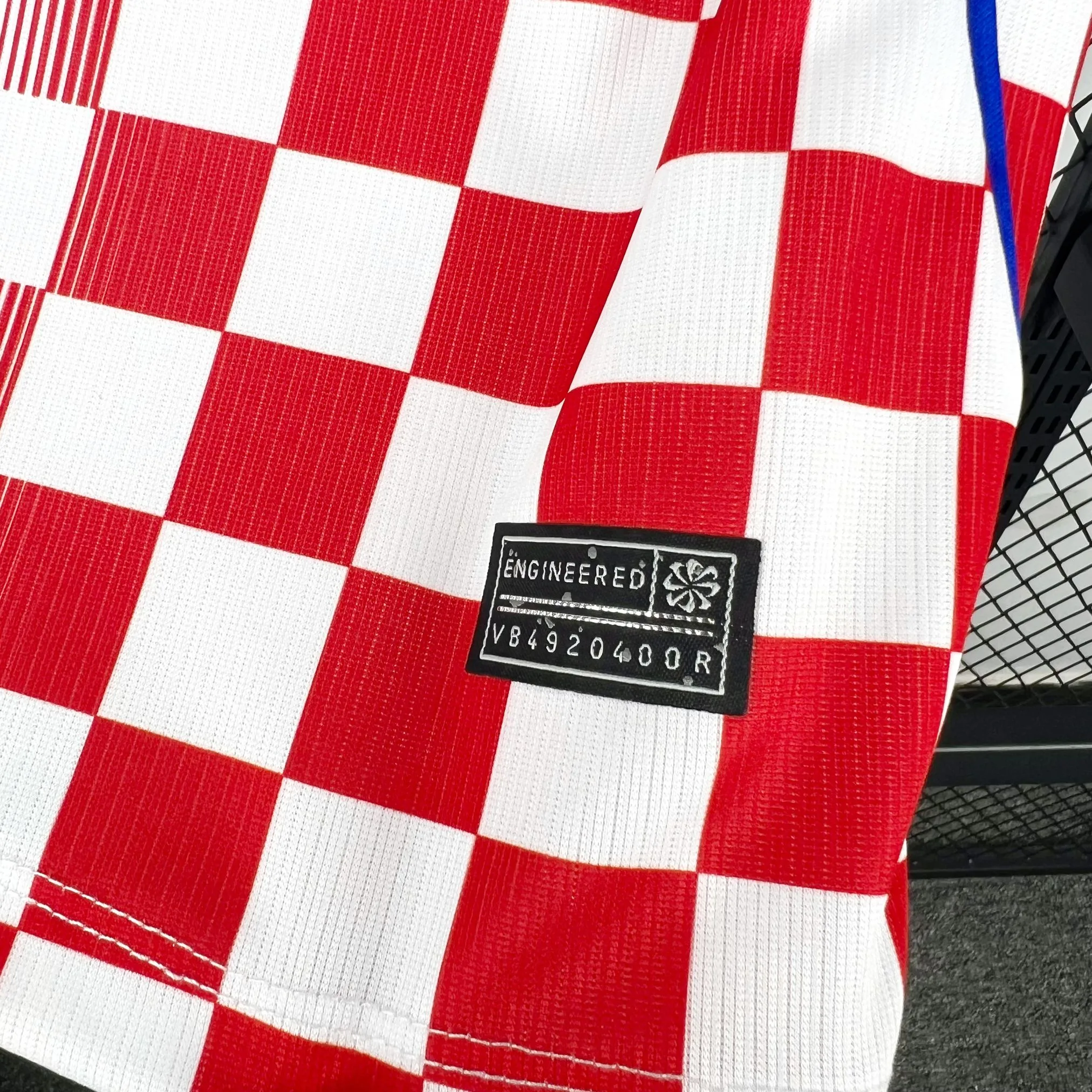 Detalle de un parche negro rectangular "ENGINEERED" con un código de serie, cosido sobre el patrón de cuadros rojos y blancos cerca del dobladillo inferior.