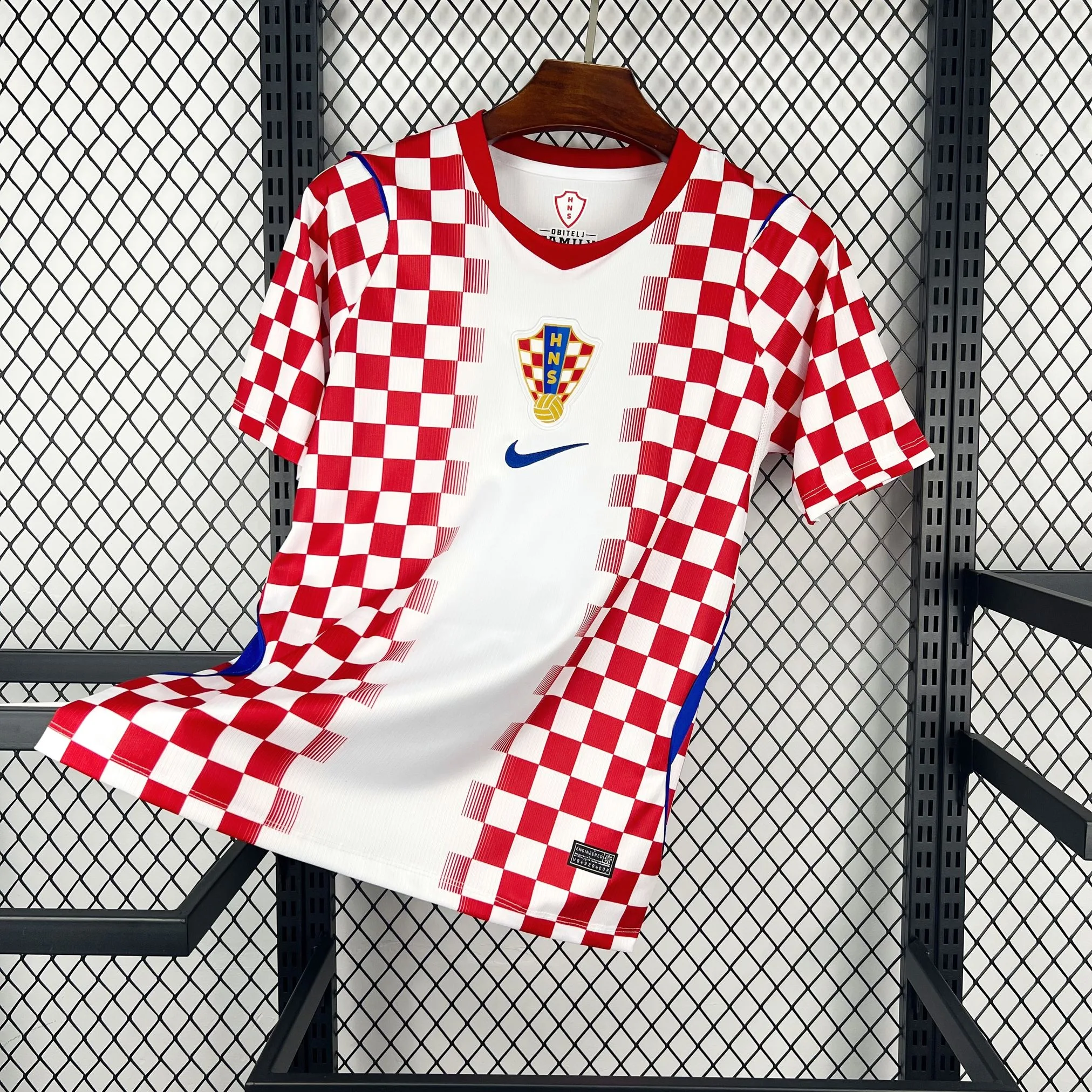 Camiseta de la selección de Croacia con su tradicional diseño de cuadros rojos y blancos ("Chequy"). El patrón se concentra en los laterales y la parte superior, dejando una franja blanca vertical en el centro donde se ubica el escudo de la federación (HNS) y el logotipo de la marca en azul. Las mangas son de color rojo sólido con detalles azules en las axilas.